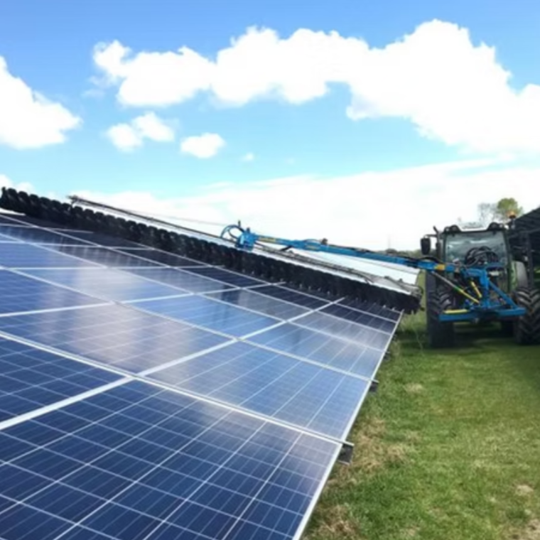 Tractor cleaning or maintaining large solar panels installed in a grassy field under a partly cloudy sky.
