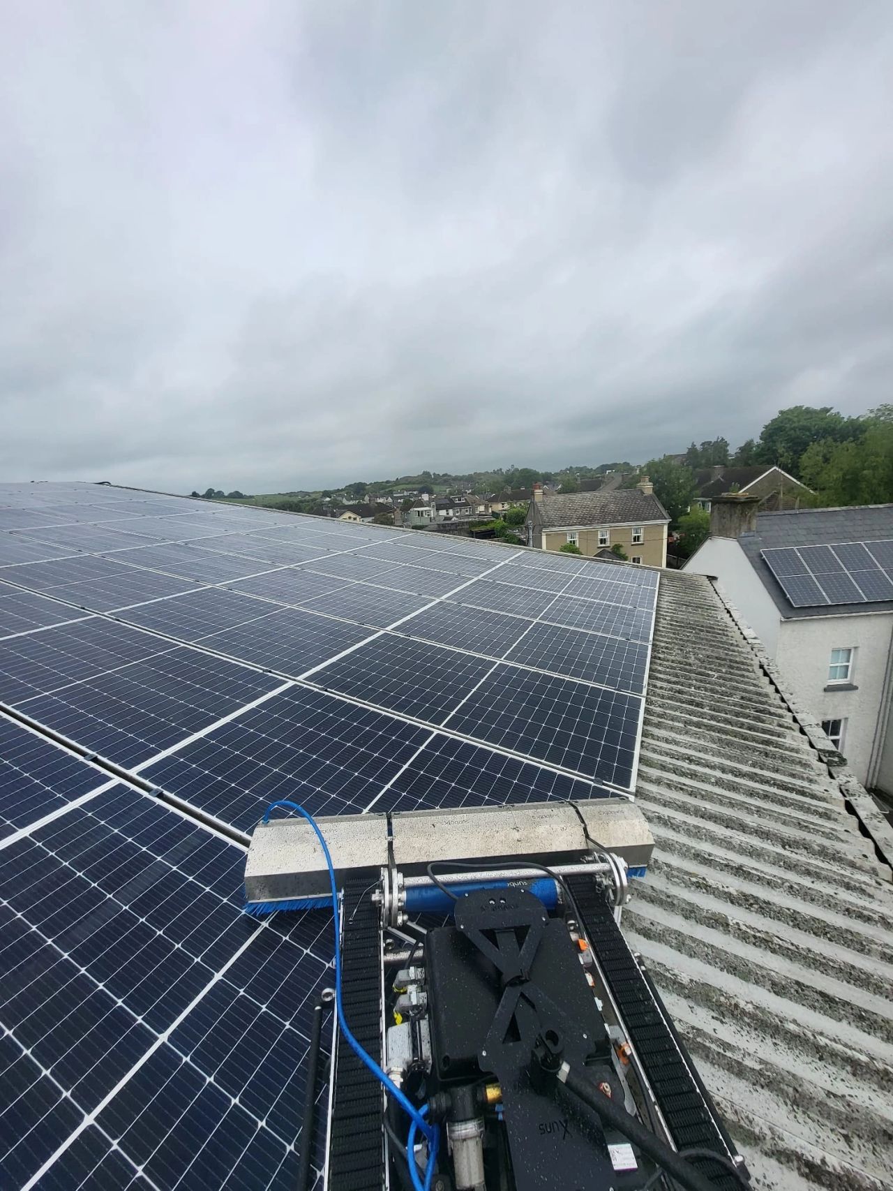 Robot cleaning large solar panels installed on a sloped roof with houses and cloudy sky in the background.