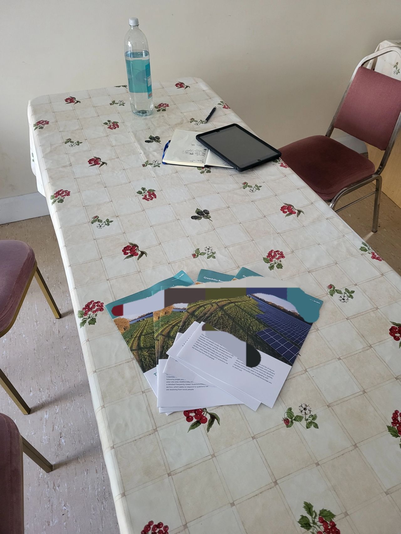 Table with a floral tablecloth holding a water bottle, an open notebook, a tablet, and several brochures with images of solar panels and farmland.