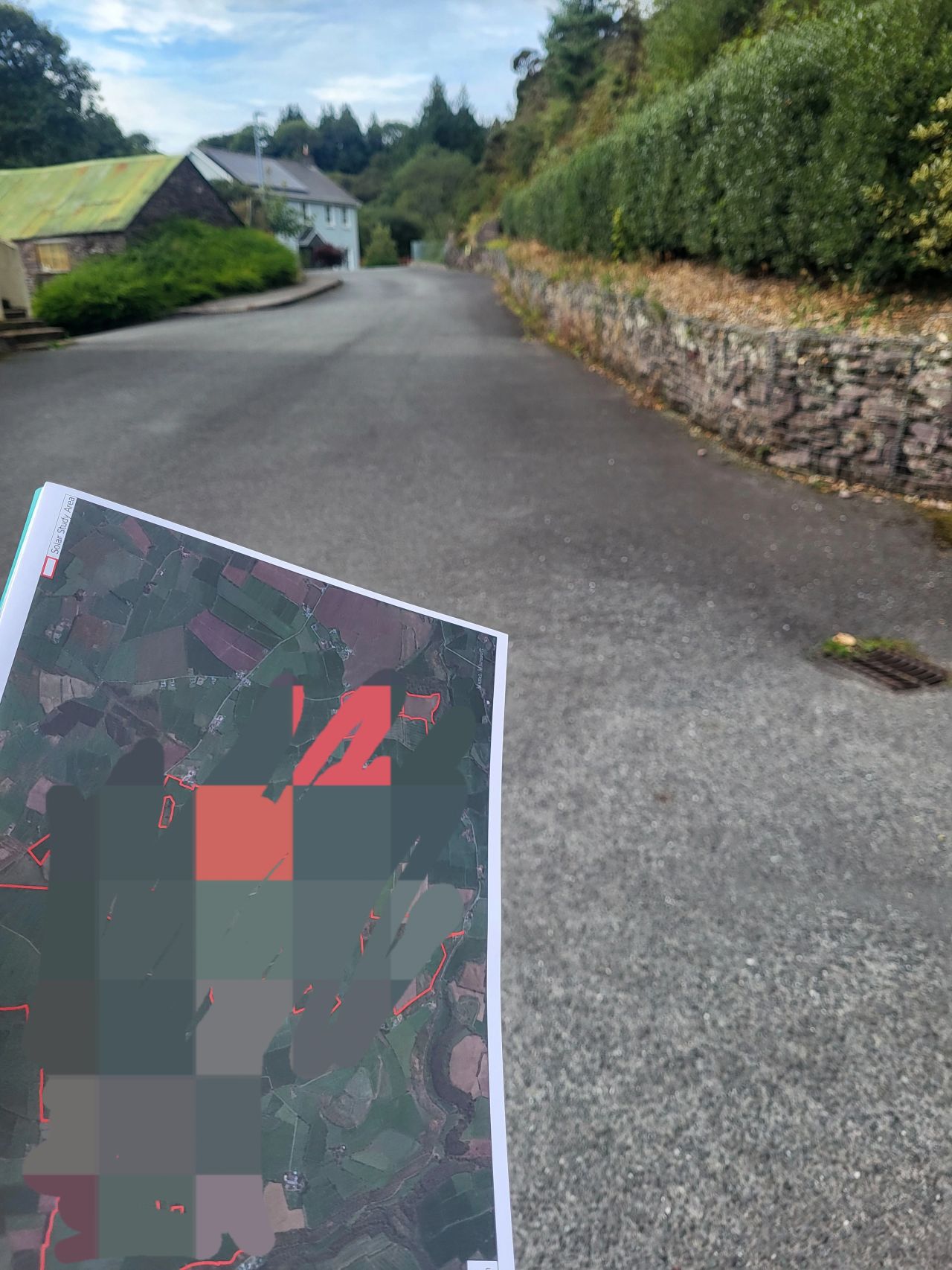 A person holding a printed map with blurred areas, standing on a quiet paved road with stone walls and greenery on the sides.