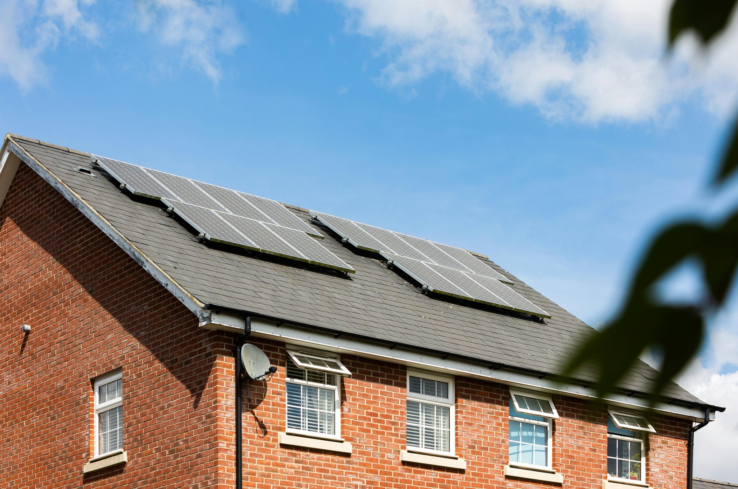Brick house with four windows and solar panels installed on the sloped roof under a partly cloudy sky.
