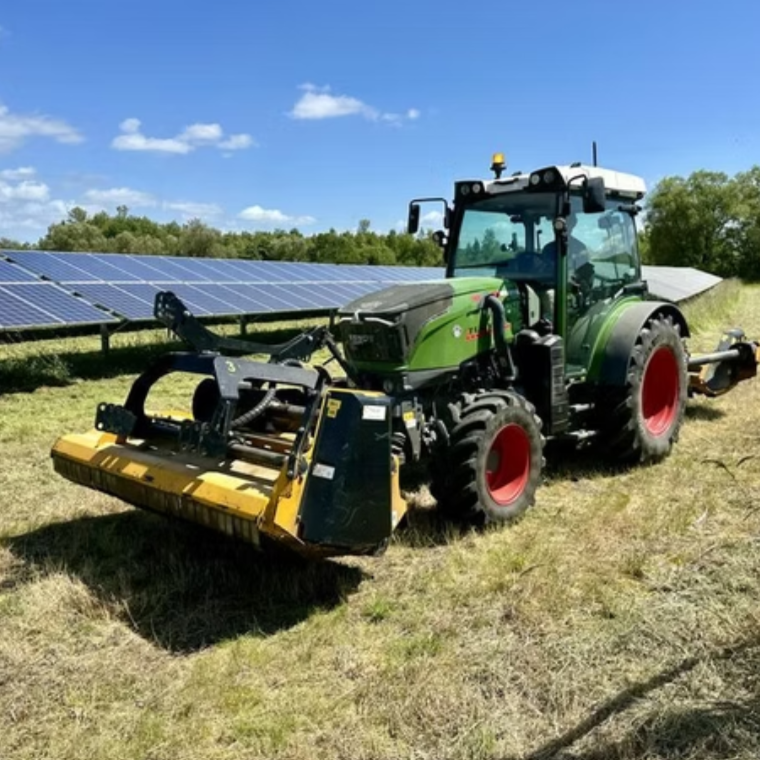 Green tractor with red wheels parked on grass next to solar panels under a blue sky.