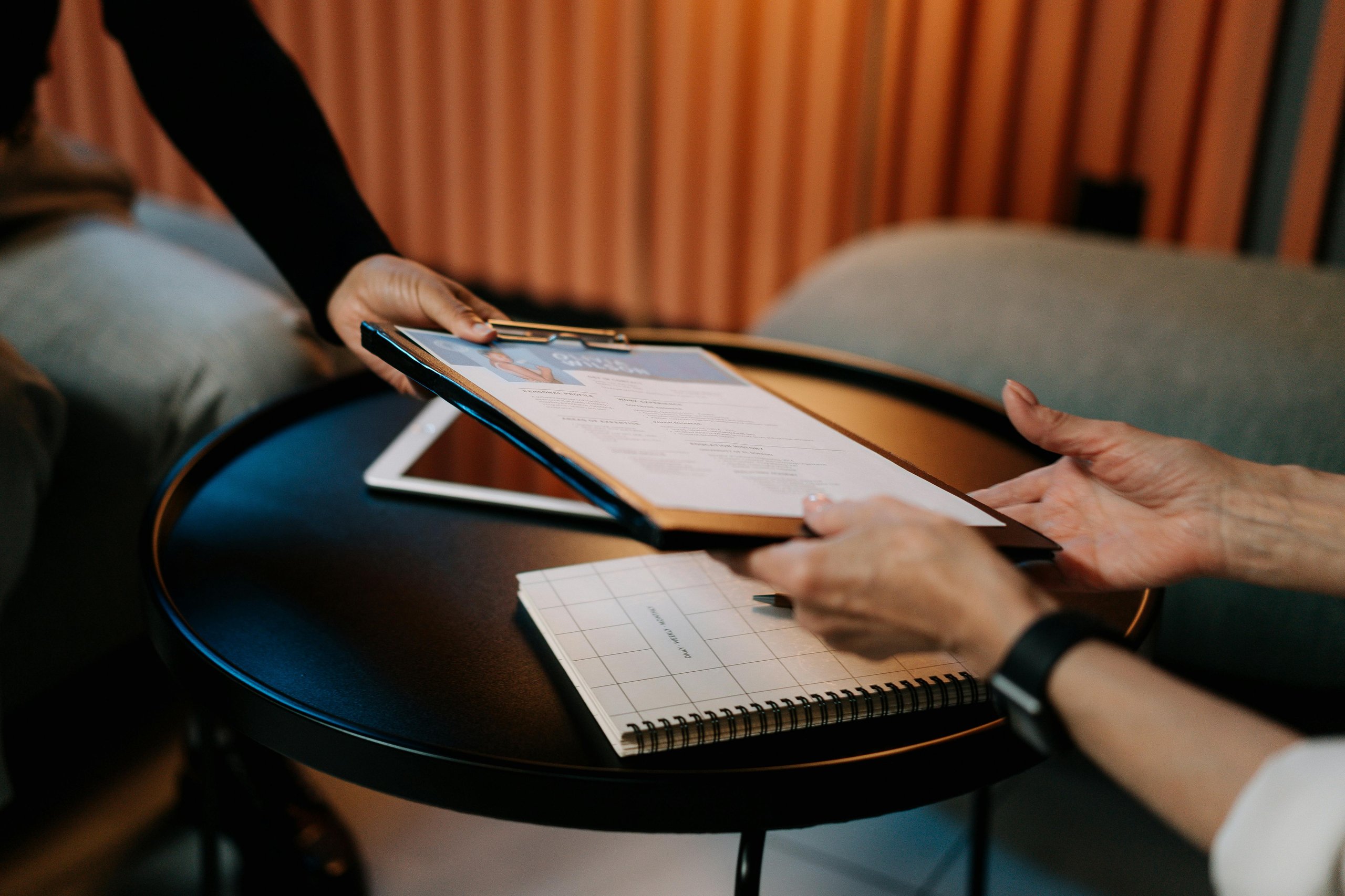 Two people exchanging a clipboard with a document over a round black table with a notebook and tablet.