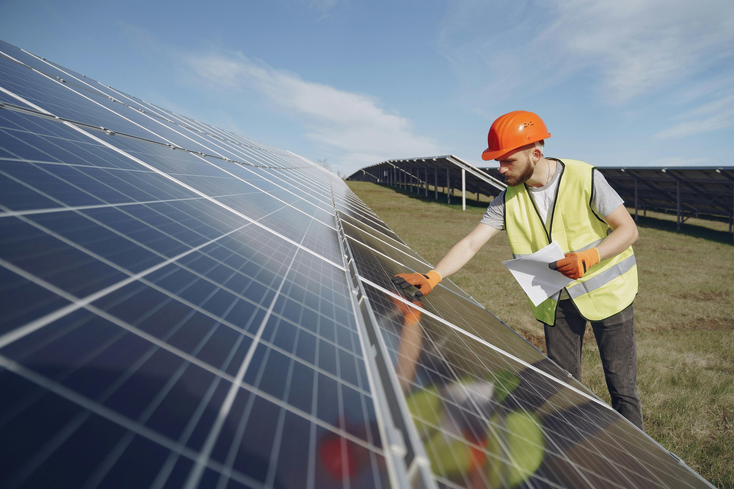 Technician in safety vest and hard hat inspecting solar panels in a field under blue sky.