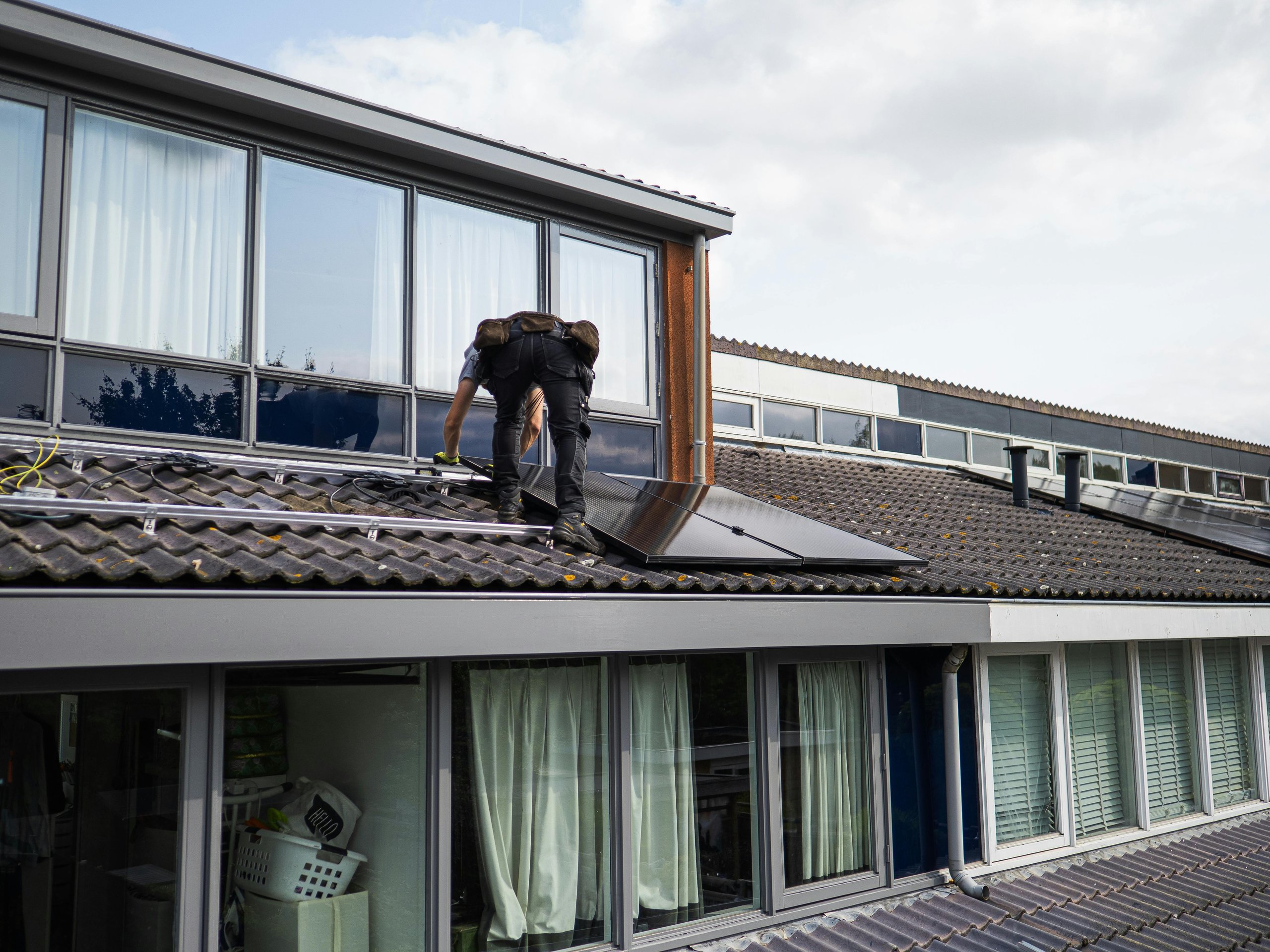 Worker installing solar panels on the tiled roof of a modern building under a cloudy sky.