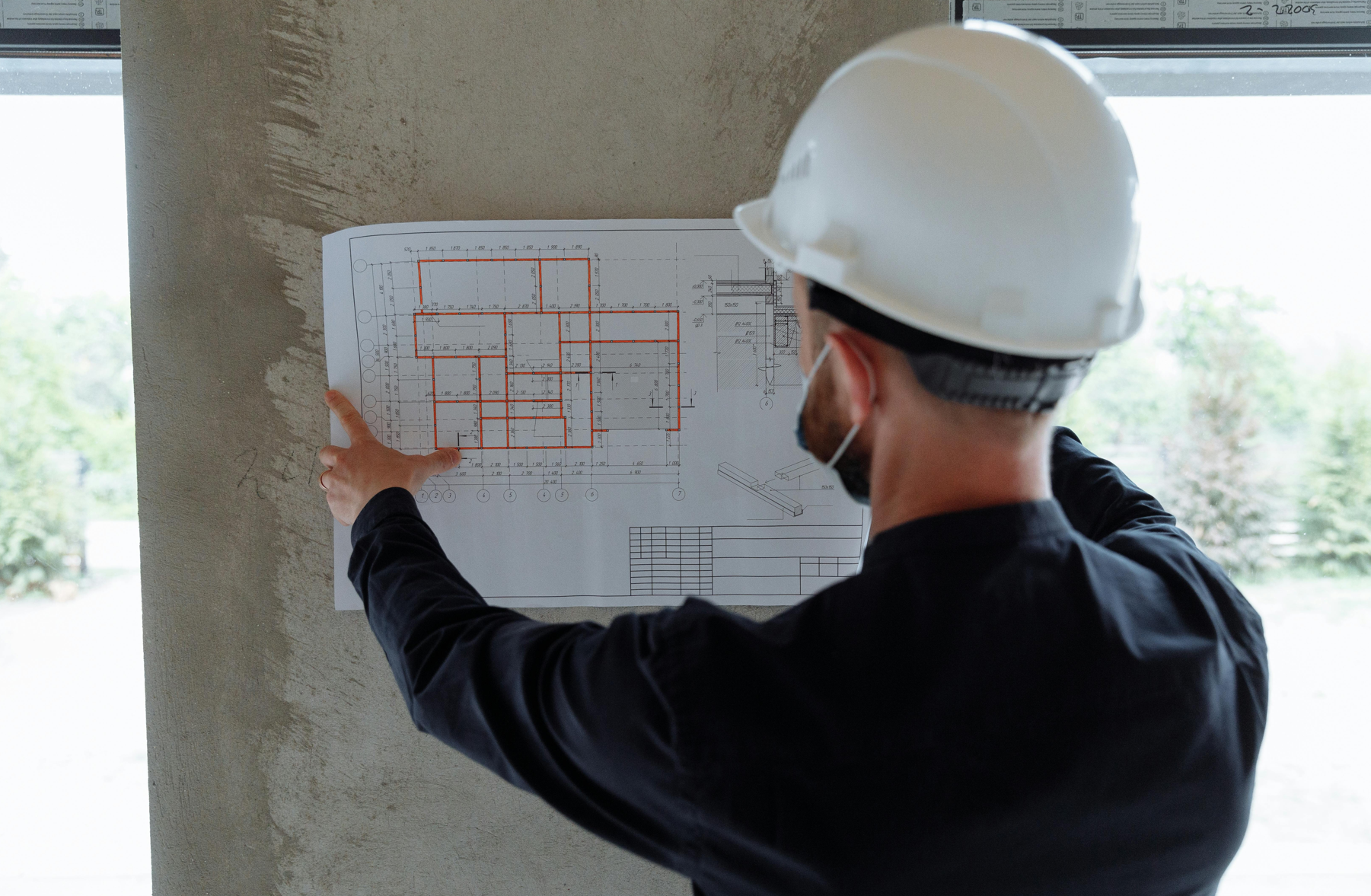 Construction worker wearing a white hard hat and dark clothing holding and reviewing a building blueprint against a concrete wall.