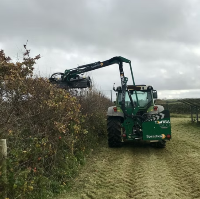 Green tractor with an attached hedge trimmer cutting tall bushes under a cloudy sky.