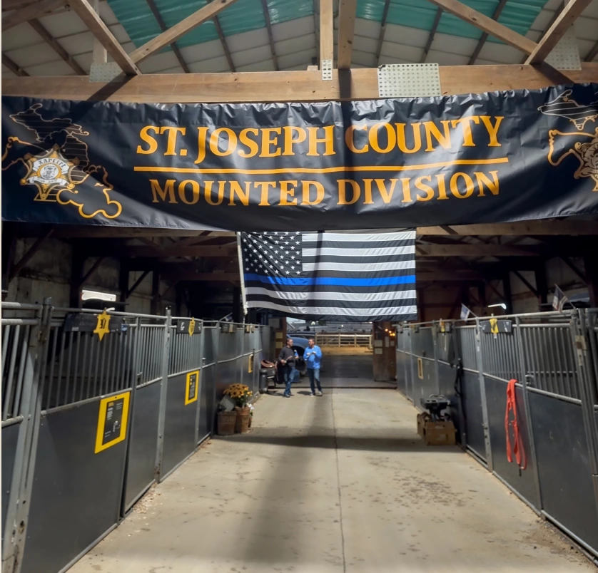 Indoor stable aisle with a banner reading 'St. Joseph County Mounted Division' hanging above and a black and white Thin Blue Line flag, with two people standing at the end.