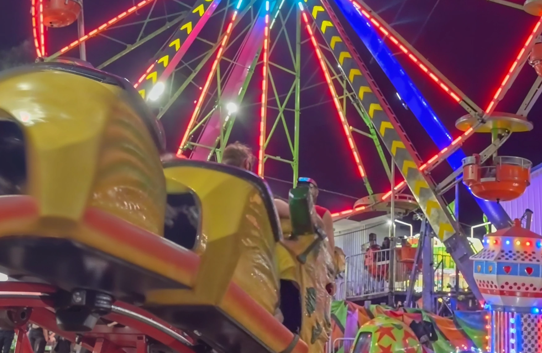 Close-up of yellow carnival ride seats with a brightly lit Ferris wheel in the background at night.