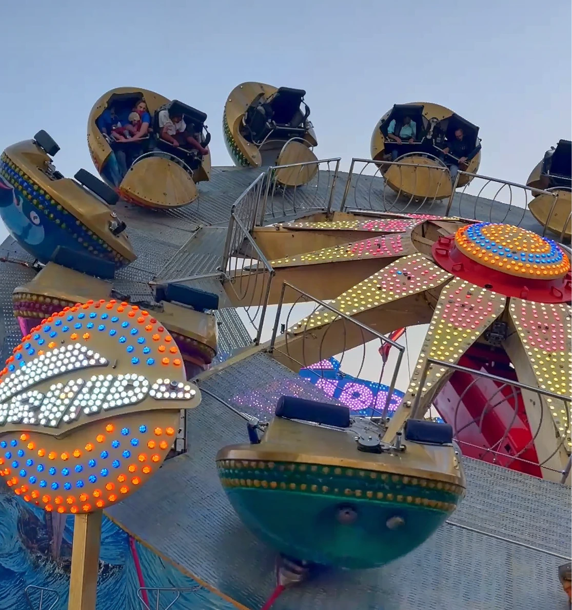 People riding a spinning carnival ride with round, colorful seats elevated against a clear sky.