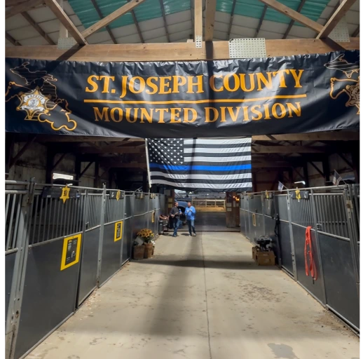 Interior of a stable with metal horse stalls on both sides, a large banner reading 'ST. JOSEPH COUNTY MOUNTED DIVISION' overhead, and a black-and-white American flag with a blue line hanging in the background.