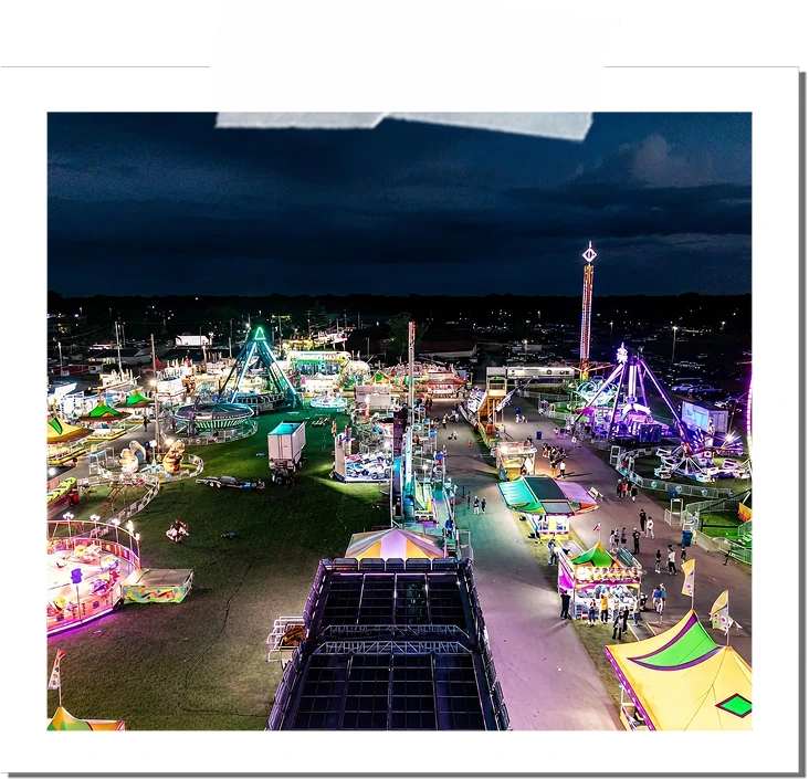 Aerial nighttime view of a brightly lit carnival with rides, booths, and people walking along pathways.