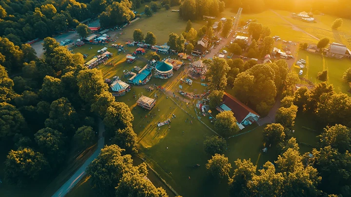 Aerial view of a small fair with tents, carnival rides, and people surrounded by trees during golden hour.