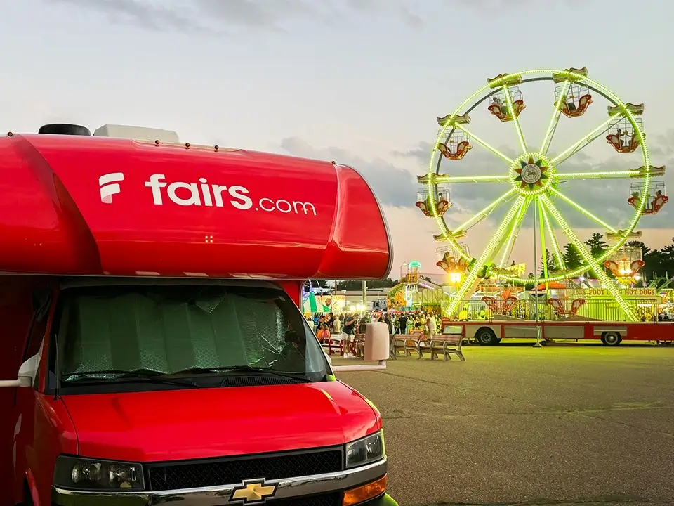Red Chevrolet RV with fairs.com logo parked near a brightly lit Ferris wheel at a fairground during dusk.