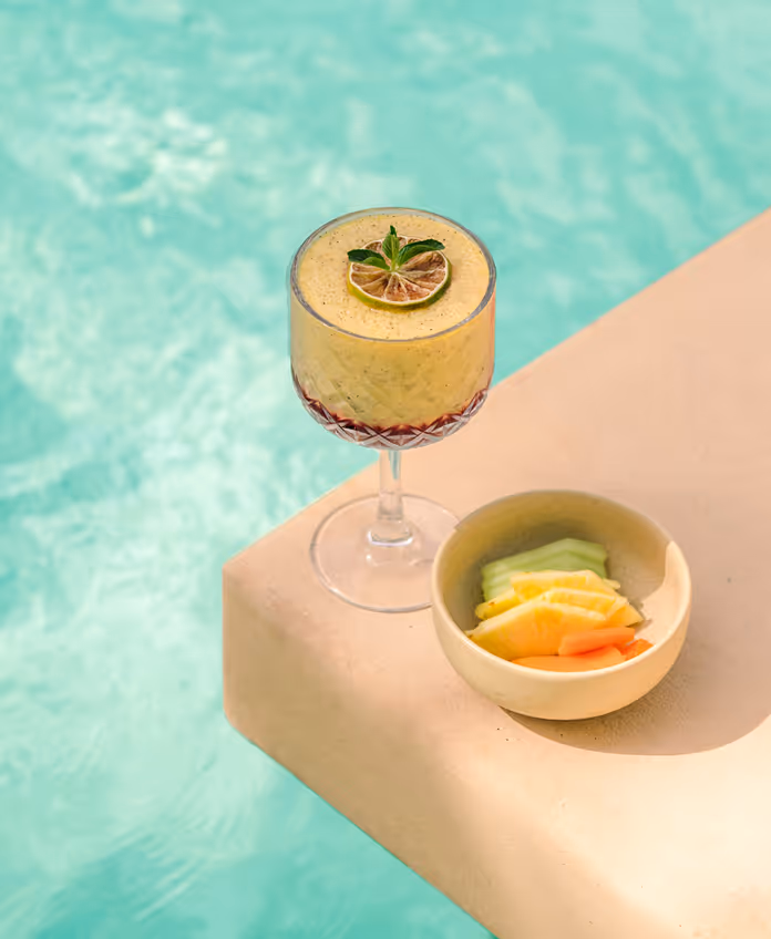 Tropical cocktail with a lime slice and a bowl of fresh fruit on the edge of the turquoise pool at La Valise Tulum hotel.