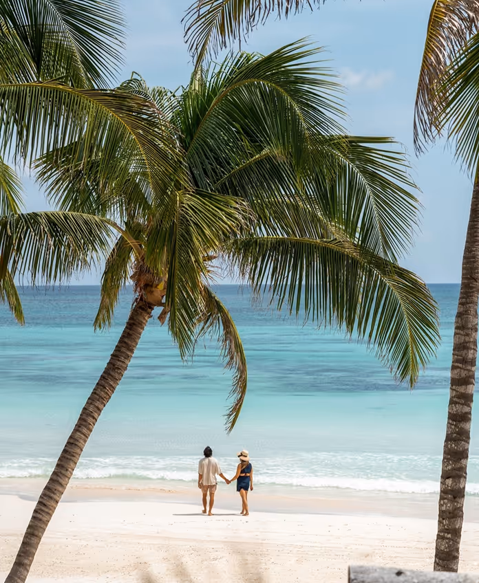 A couple walking hand in hand on the white sand beach in front of La Valise Tulum hotel, framed by tropical palm trees and turquoise Caribbean water.