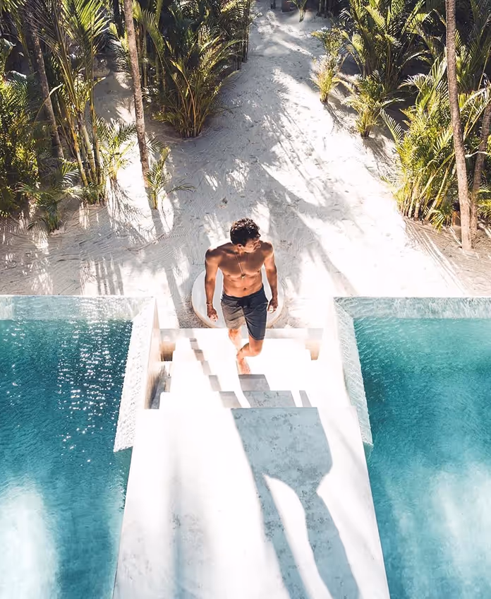 Aerial view of a man walking down the white stairs of the infinity pool at La Valise Tulum hotel, surrounded by palm trees and white sand in Mexico.