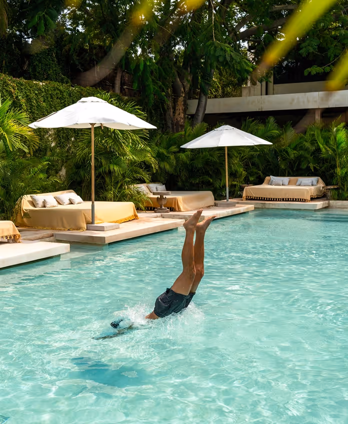 A man diving into the turquoise swimming pool at La Valise Tulum hotel, featuring luxury daybeds, white umbrellas, and lush tropical greenery.