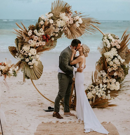 La novia y el novio se besan frente a un arco floral en la playa del hotel La Valise Tulum.