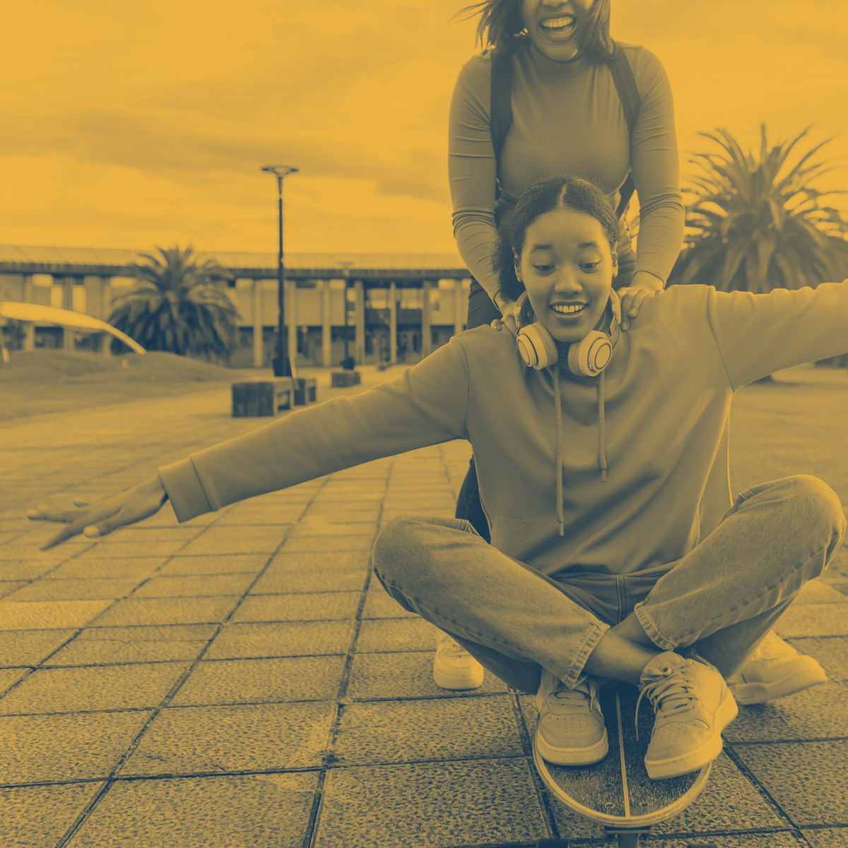 Two young women outdoors with one sitting on a skateboard and the other standing behind, holding her shoulders and smiling.