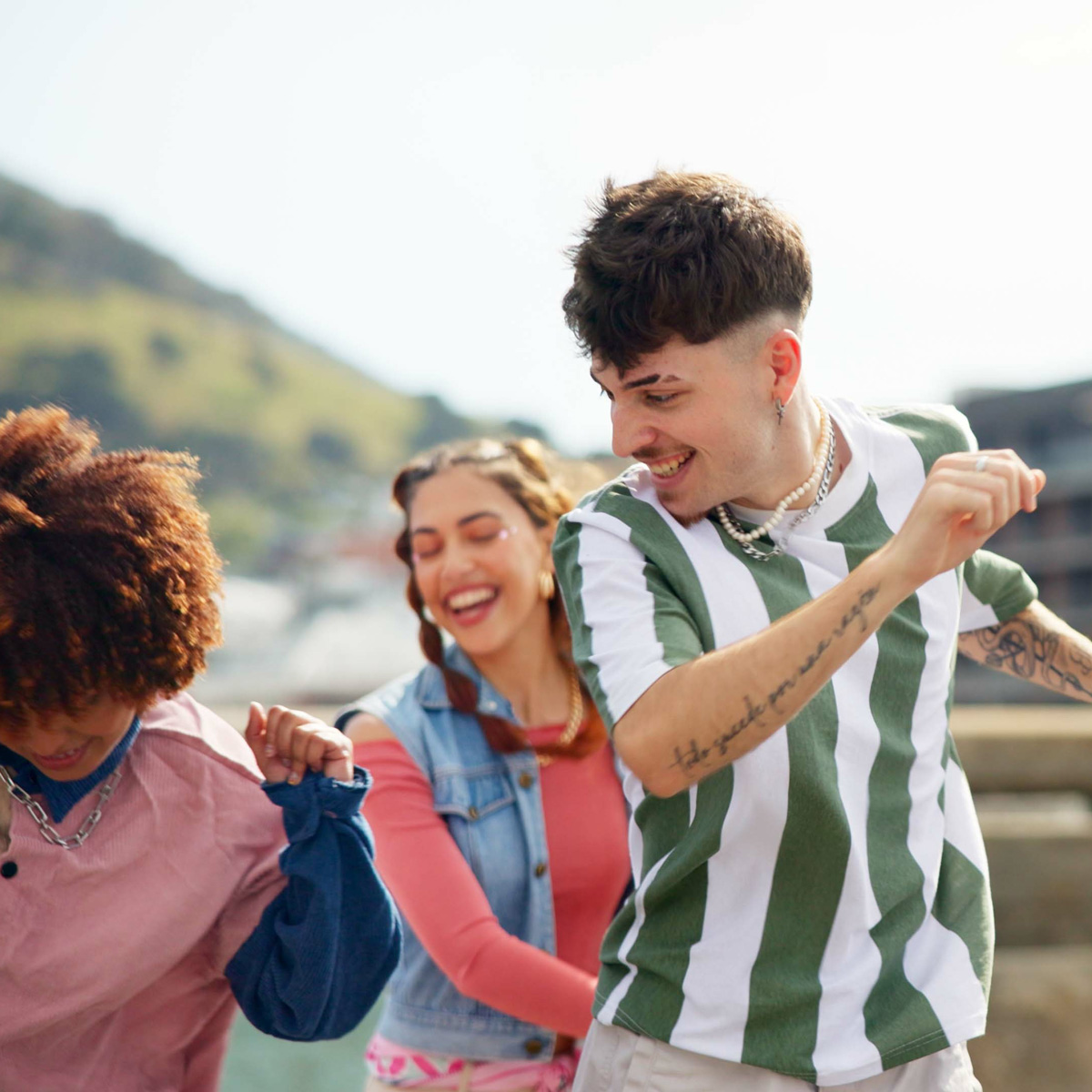 Three young adults joyfully dancing outdoors with blurred hills in the background.
