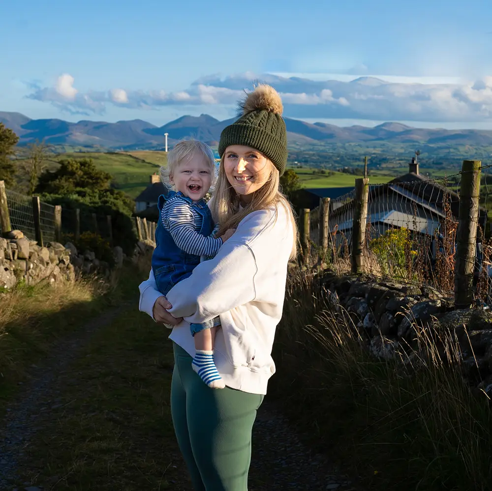 Young woman wearing a green knit hat holding a smiling toddler on a rural path with hills in the background.
