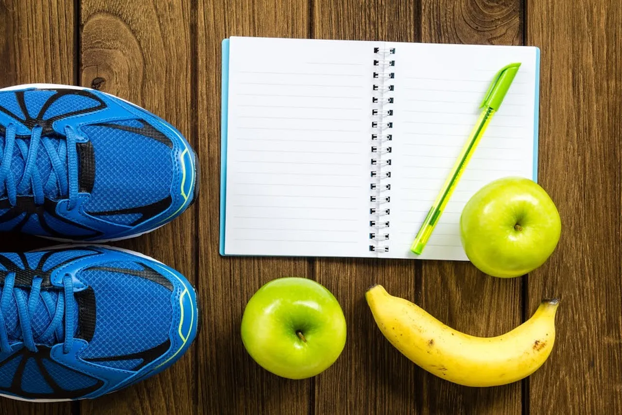 Athletic shoes next to an open notebook with a pen, surrounded by apples and bananas