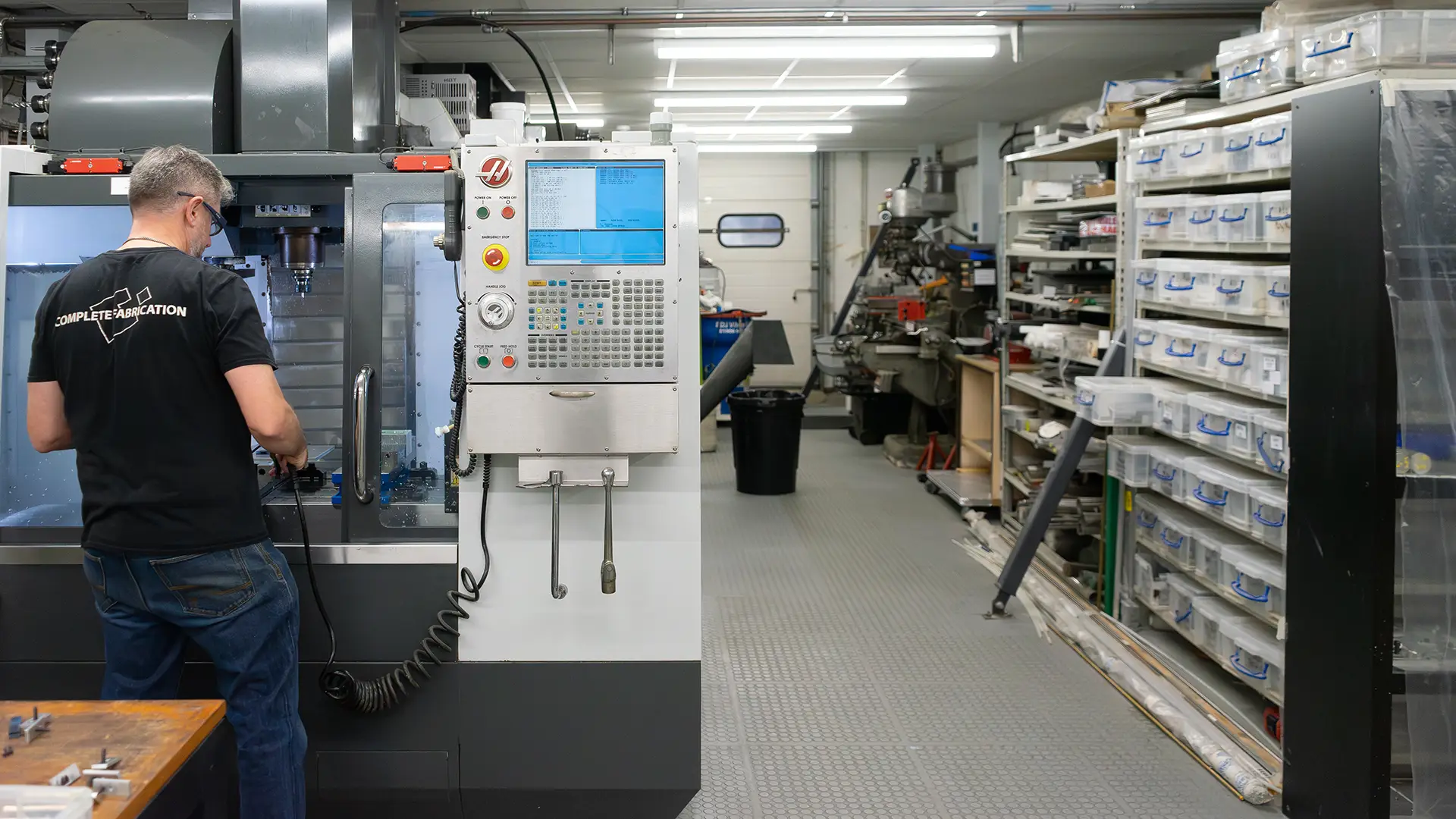 Man operating CNC machining center in a workshop with organized shelves of parts in plastic bins on the right.