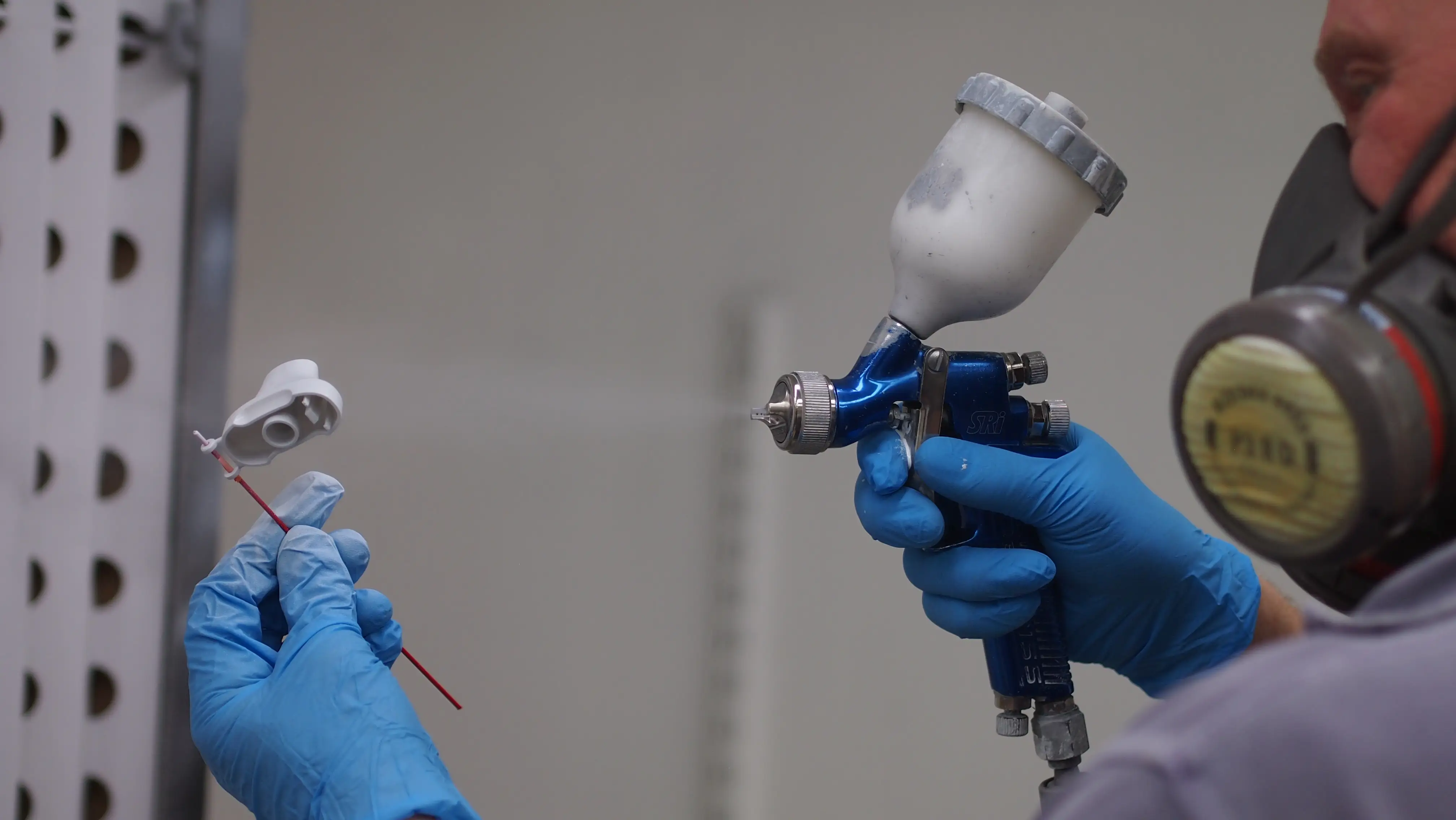Person wearing blue gloves and a respirator spray painting a small object with a blue spray gun.
