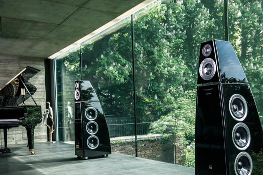 Modern interior with large floor-to-ceiling windows showing lush green trees outside, a black grand piano, and two tall black speakers.