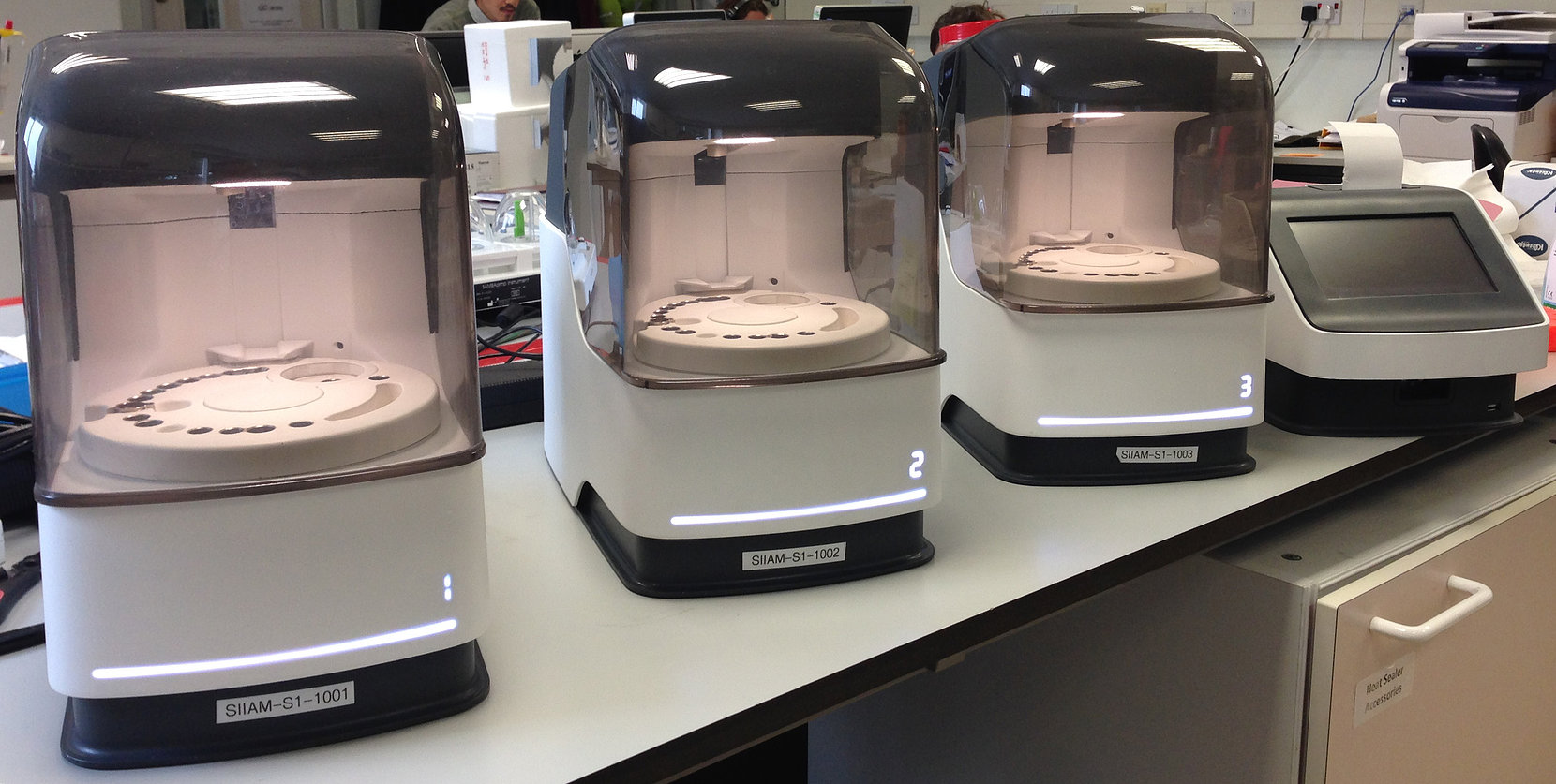 Three laboratory machines with transparent covers and circular trays numbered 1, 2, and 3 on a lab counter.