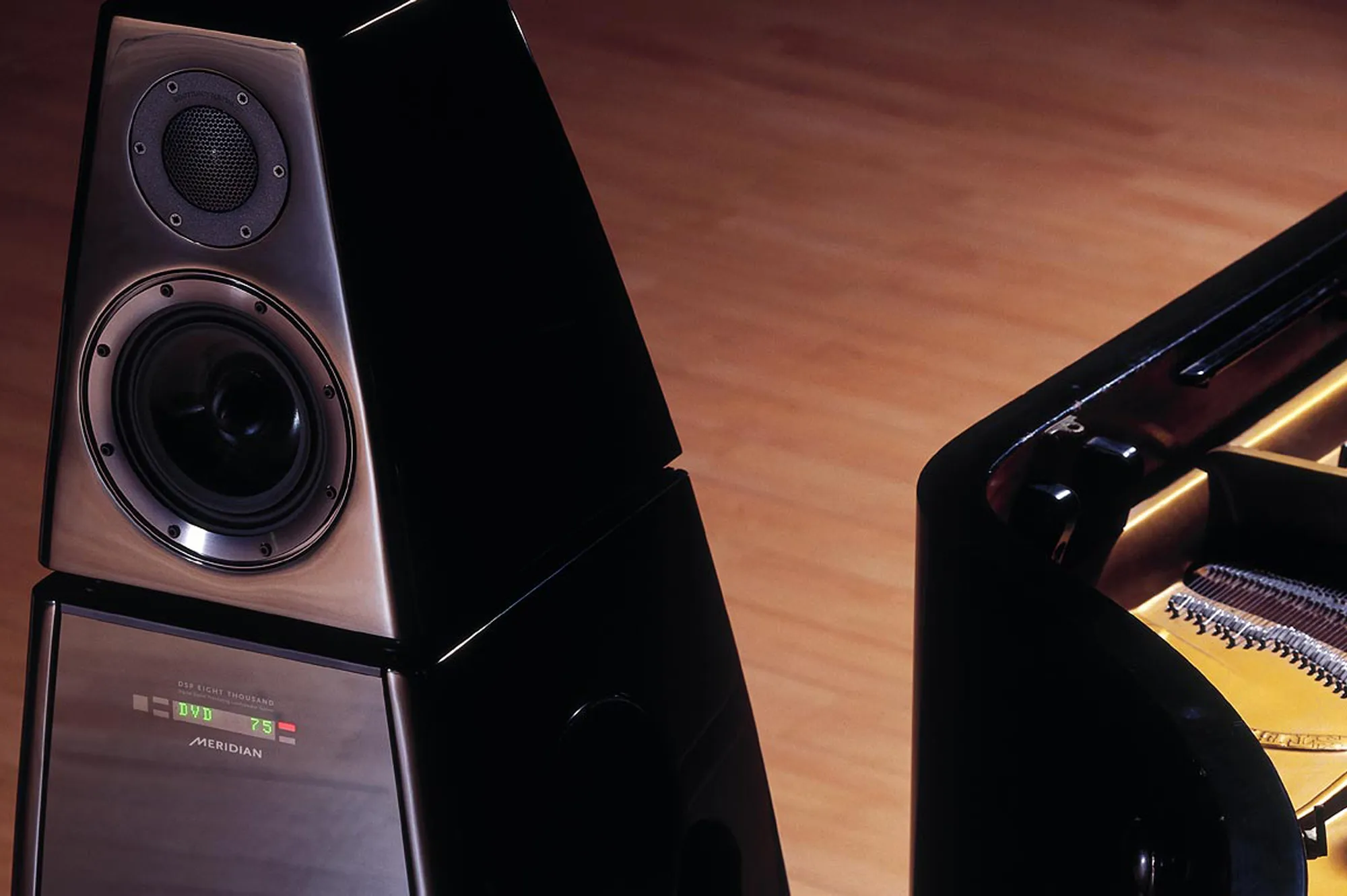 Close-up of a black Meridian DSP Eight Thousand speaker next to a grand piano showing part of its strings and tuning pins.