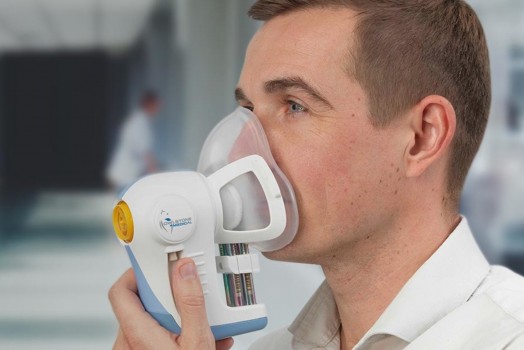 A man wearing a clear mask connected to a handheld respiratory testing device.