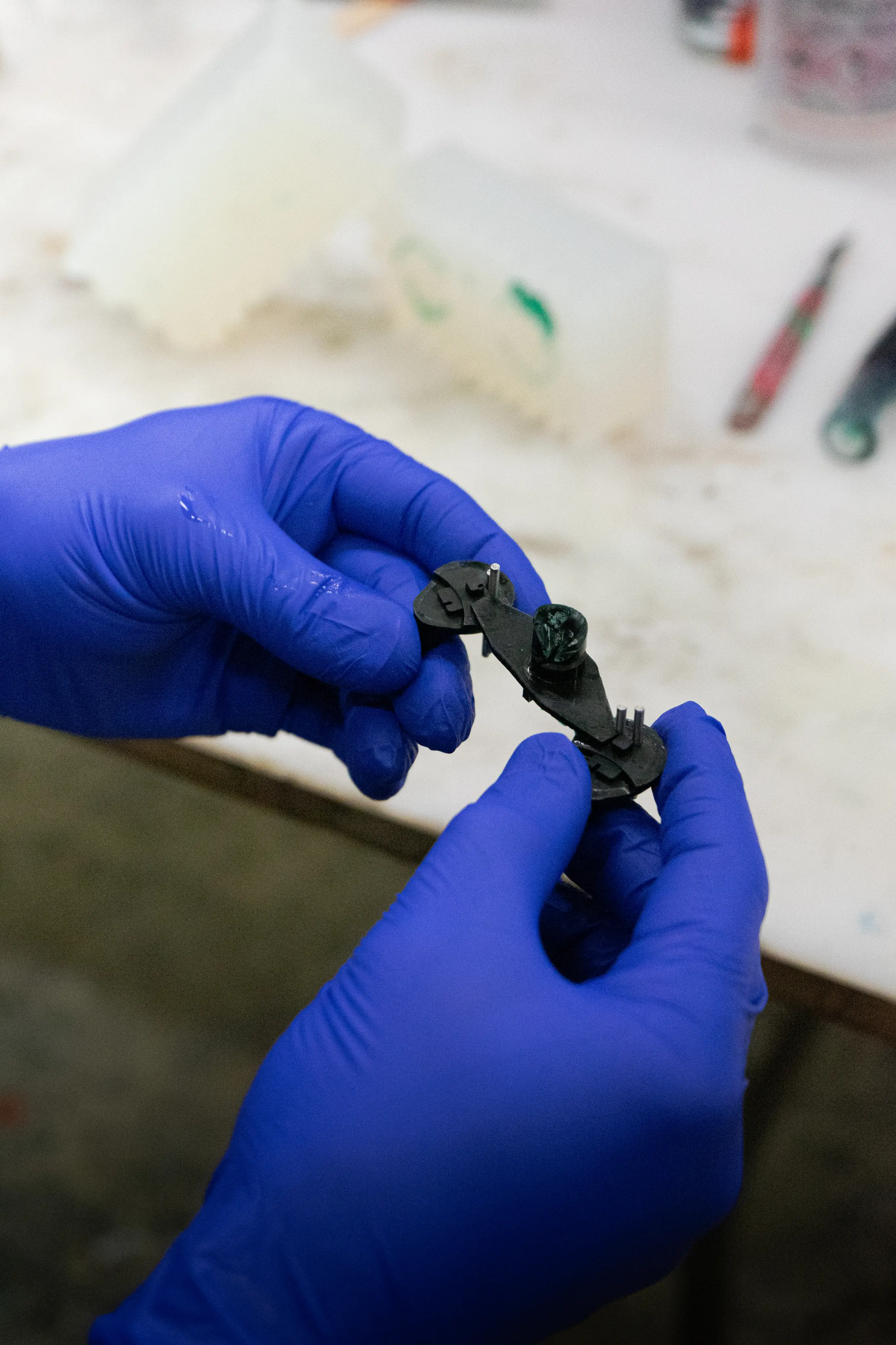 Person wearing blue gloves holding a small black plastic component with metal pins and a green-marked part.