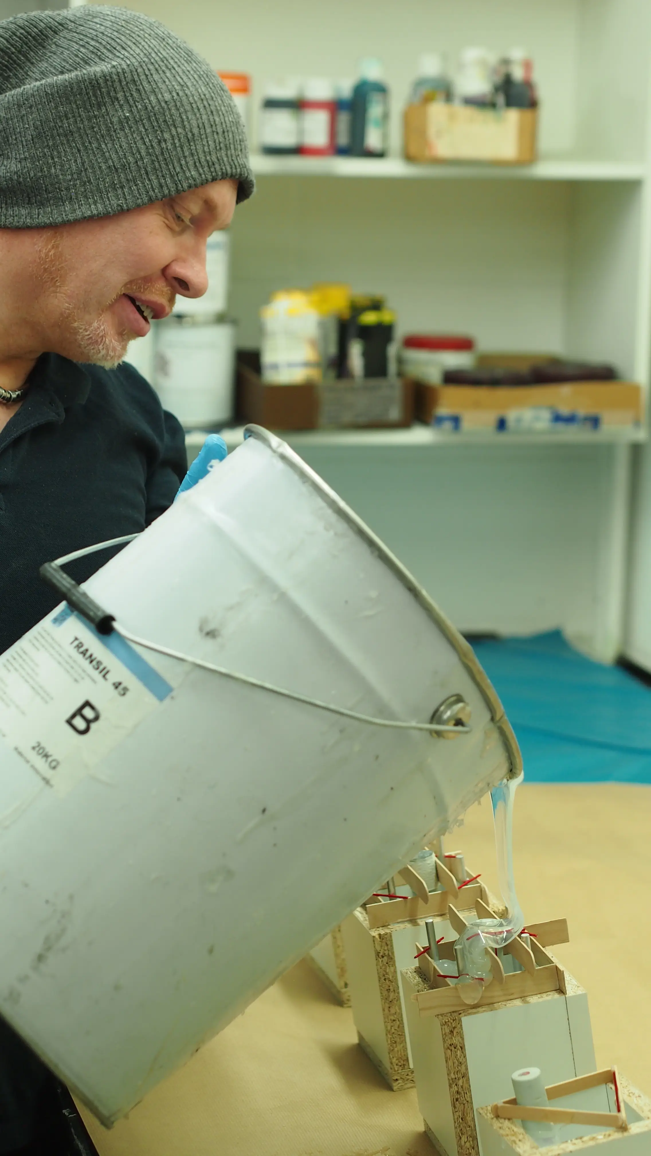 Man wearing gray beanie pouring clear liquid from a large white bucket labeled 'TRANSIL 45 B' into small containers with wooden braces on a table.