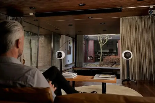 Man sitting on a couch facing a large window with two modern speakers on stands beside a coffee table.