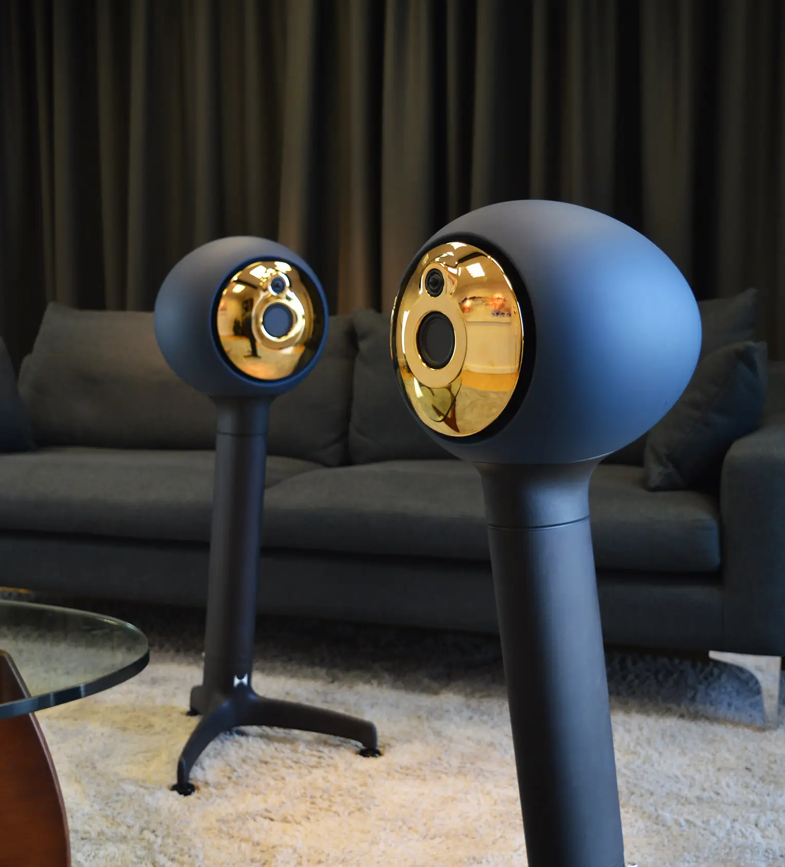 Two modern black speakers with gold reflective front panels standing on a beige carpet in front of a dark sofa with cushions.