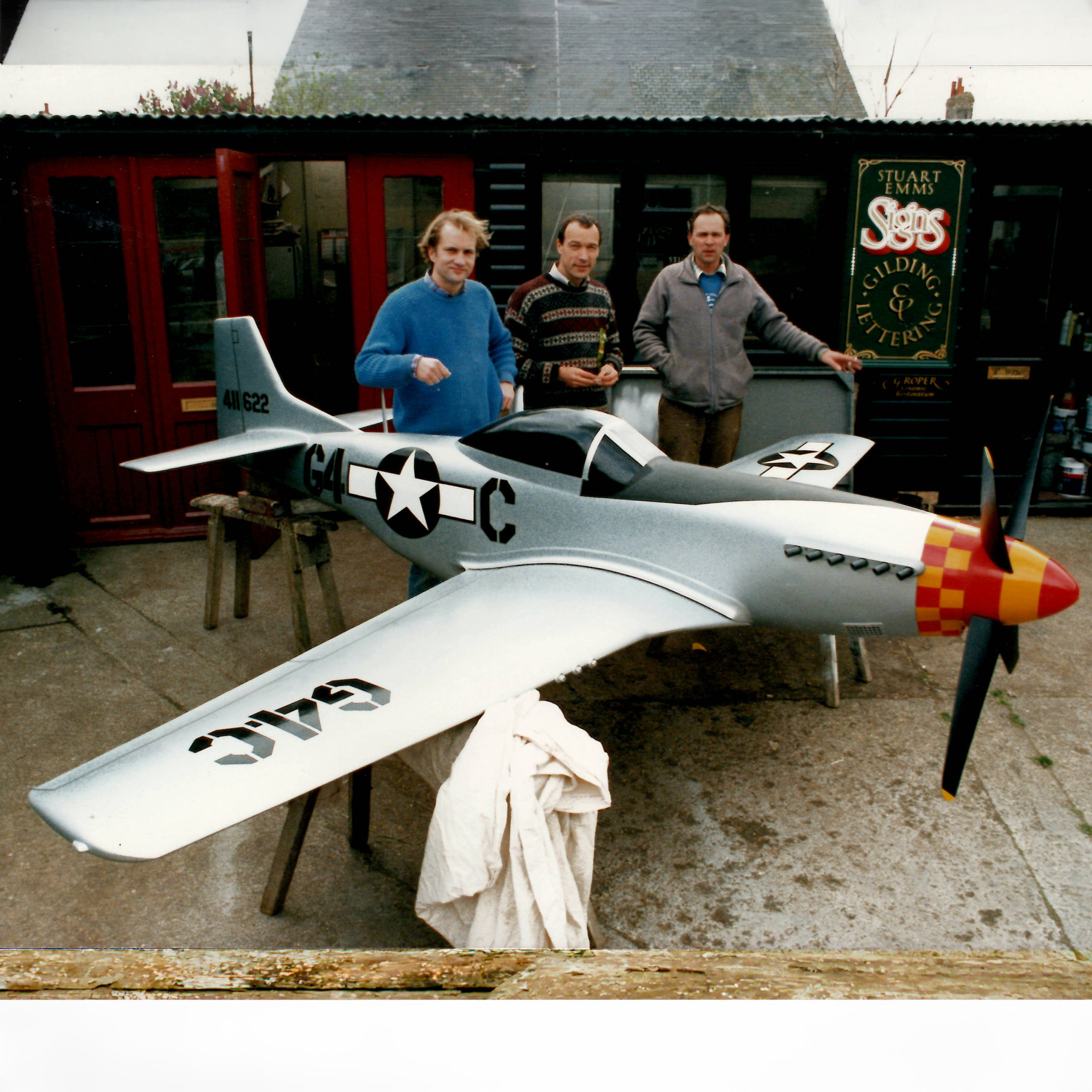 Three men standing behind a large, detailed scale model of a P-51 Mustang aircraft with U.S. Air Force insignia and a red and yellow checkered nose.