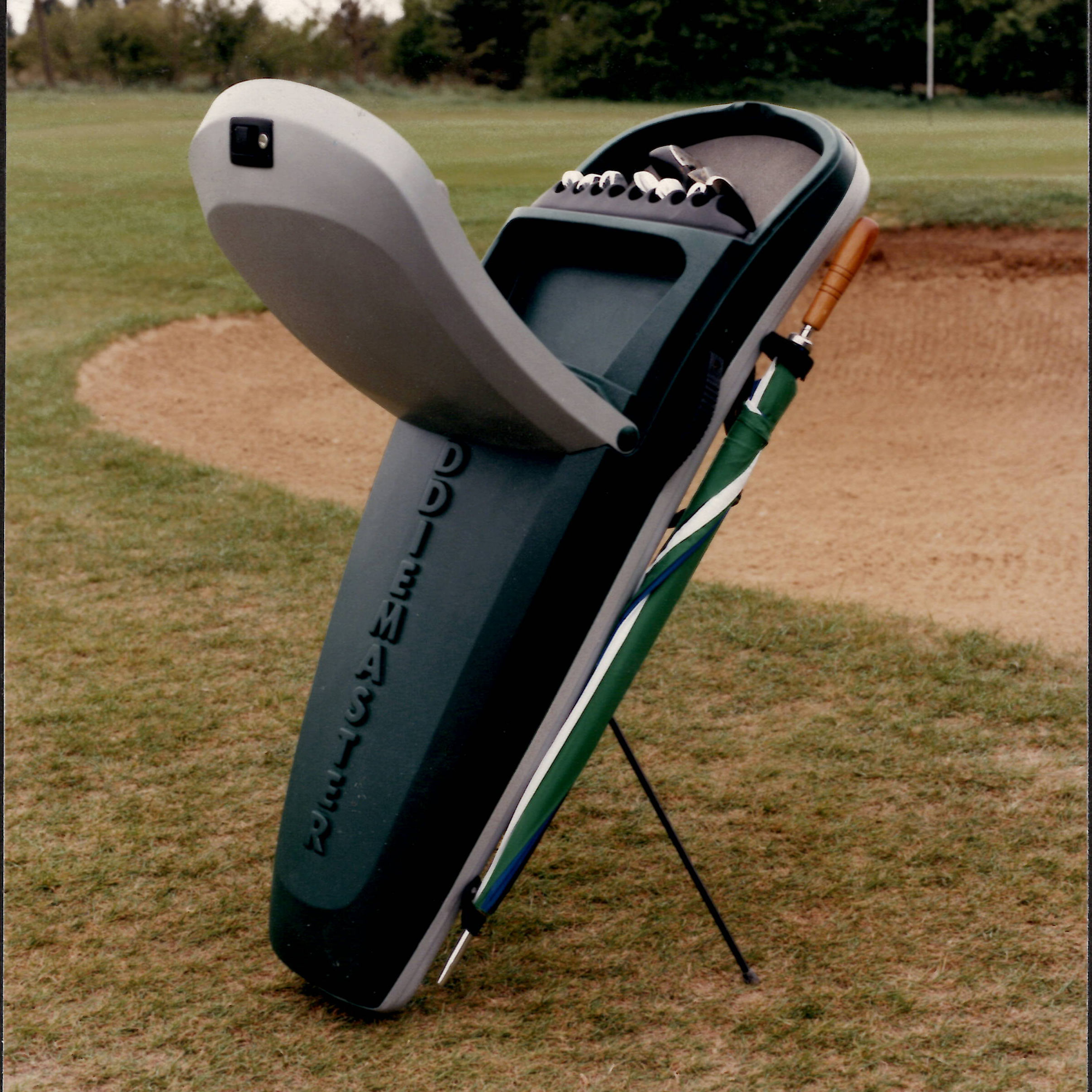 A standing golf bag with an open top cover showing golf tees and a putter on a golf course near a sand bunker, with an umbrella clipped to the side leg support.