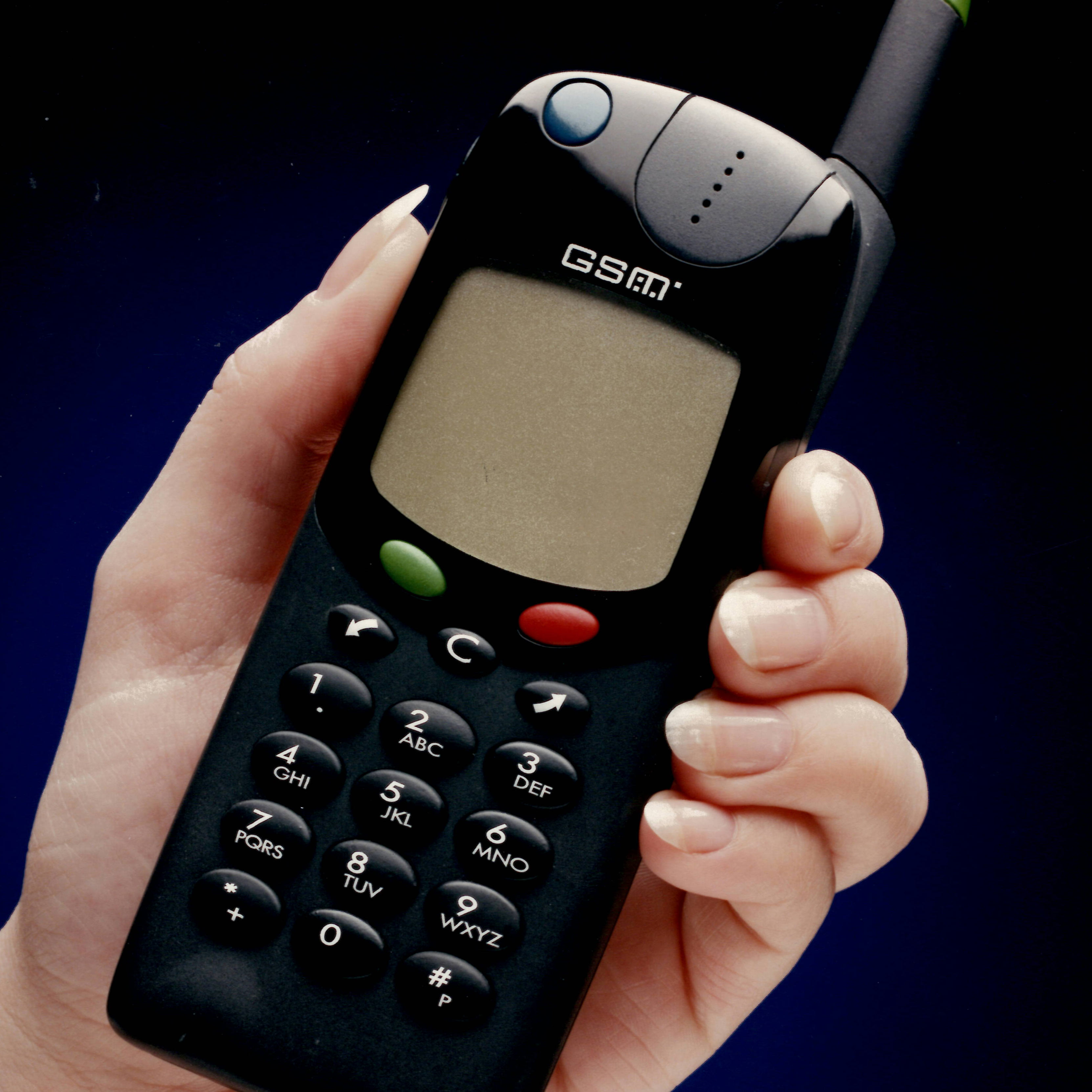 Hand holding an old black GSM mobile phone with a small monochrome screen, physical numeric keypad with letters, green and red buttons, and an antenna.