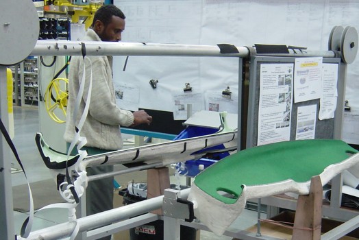 Two-operator assembly jig with green baize-lined nest and tape spools on a dashboard production line