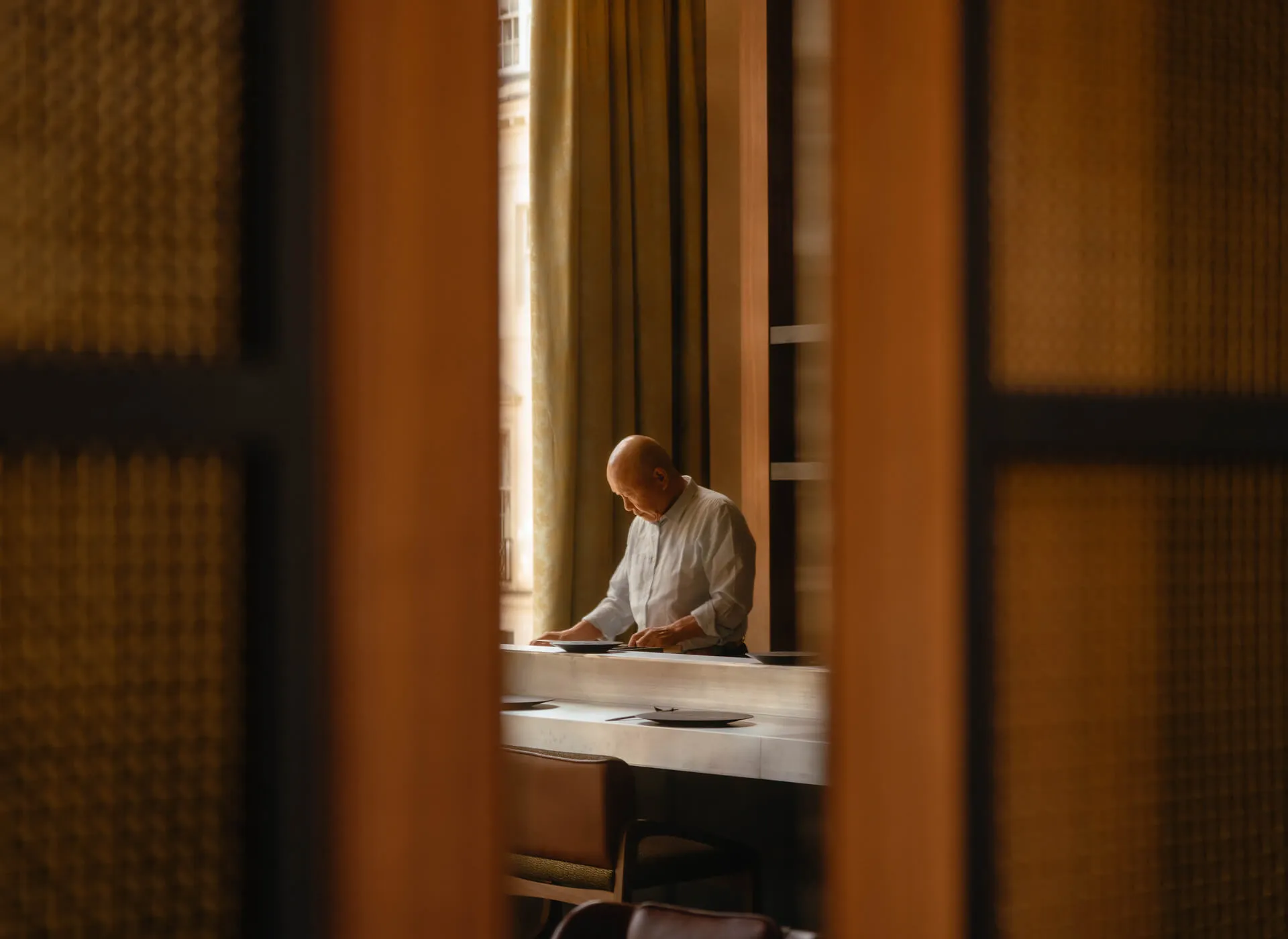 Focus shot of Chef Masa Takayama through some meshed sliding doors setting some table-wear. 