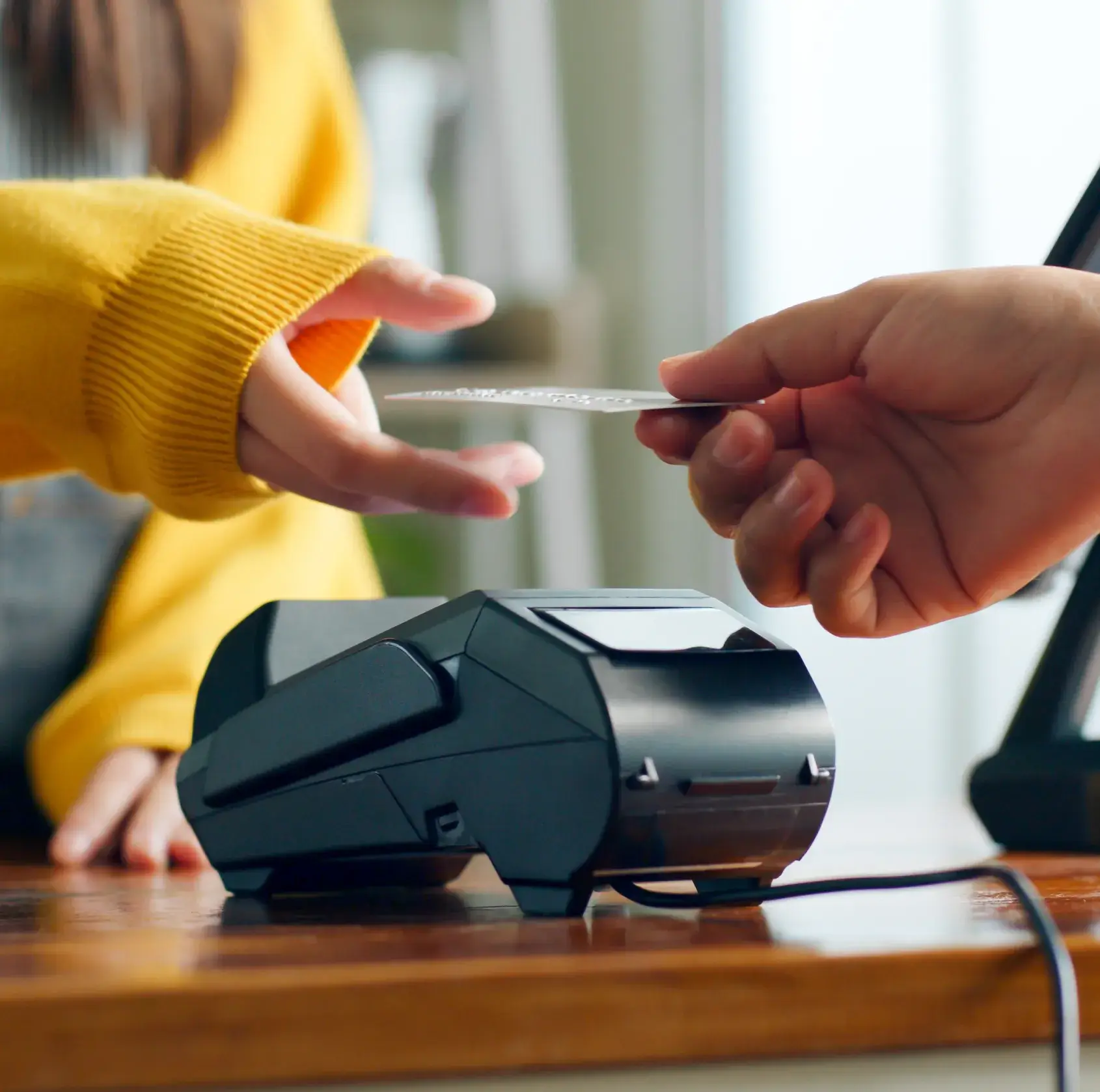 Person in a yellow sweater about to swipe a credit card on a black card reader at a wooden counter.