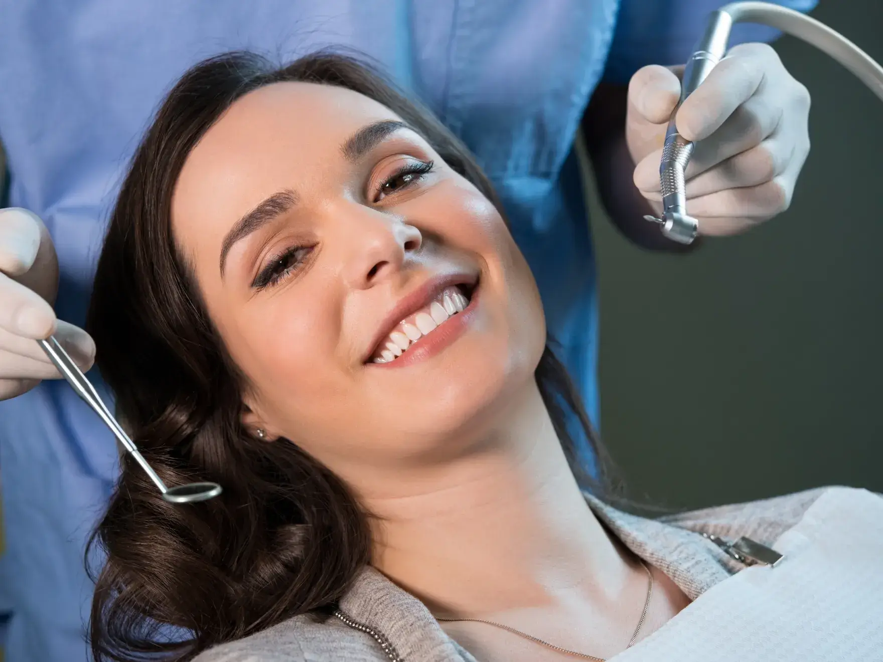 Smiling woman reclining in a dental chair with dentist’s gloved hands holding dental tools near her mouth.
