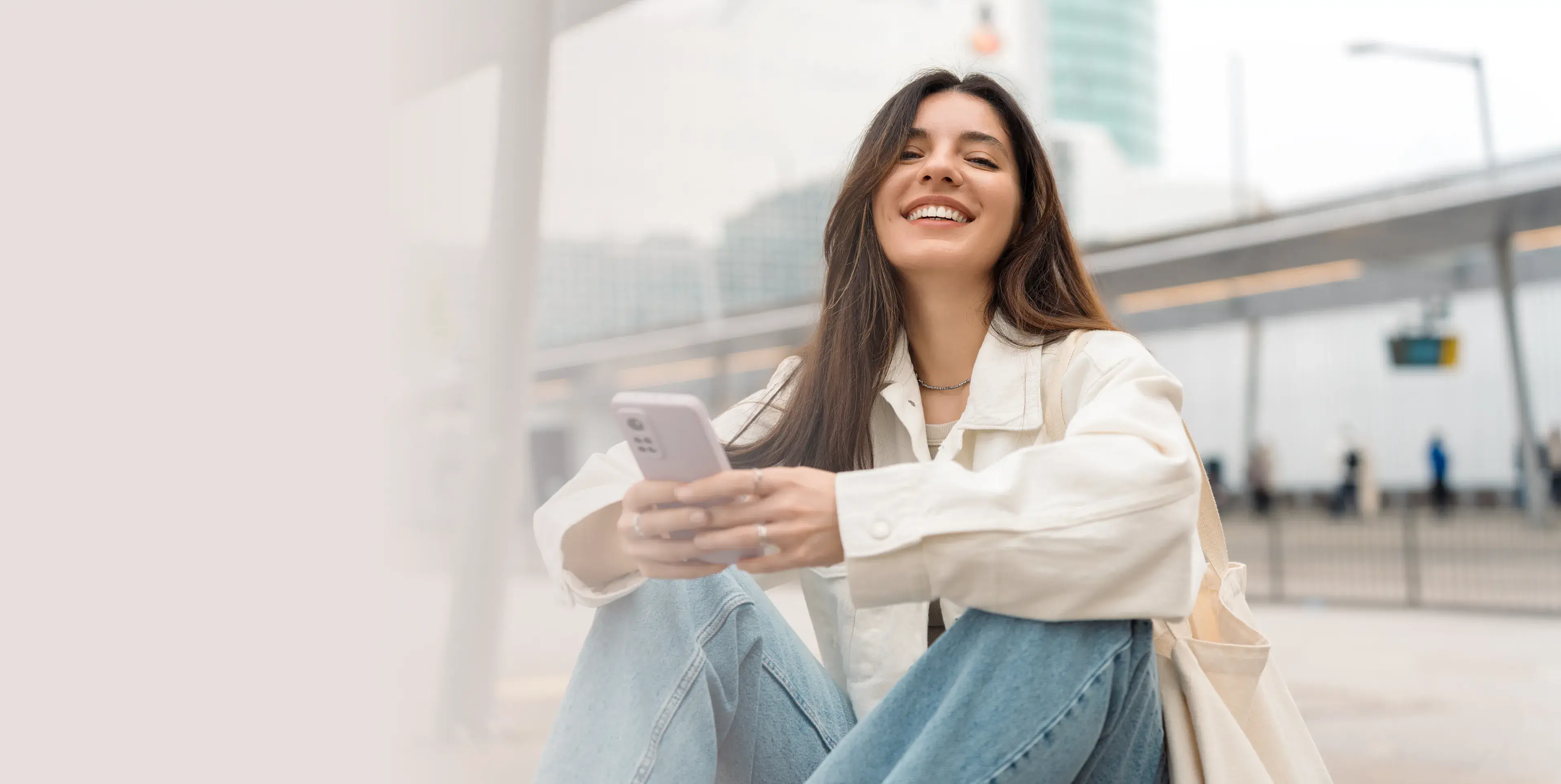 Smiling young woman in a white jacket and jeans sitting outdoors using a smartphone.
