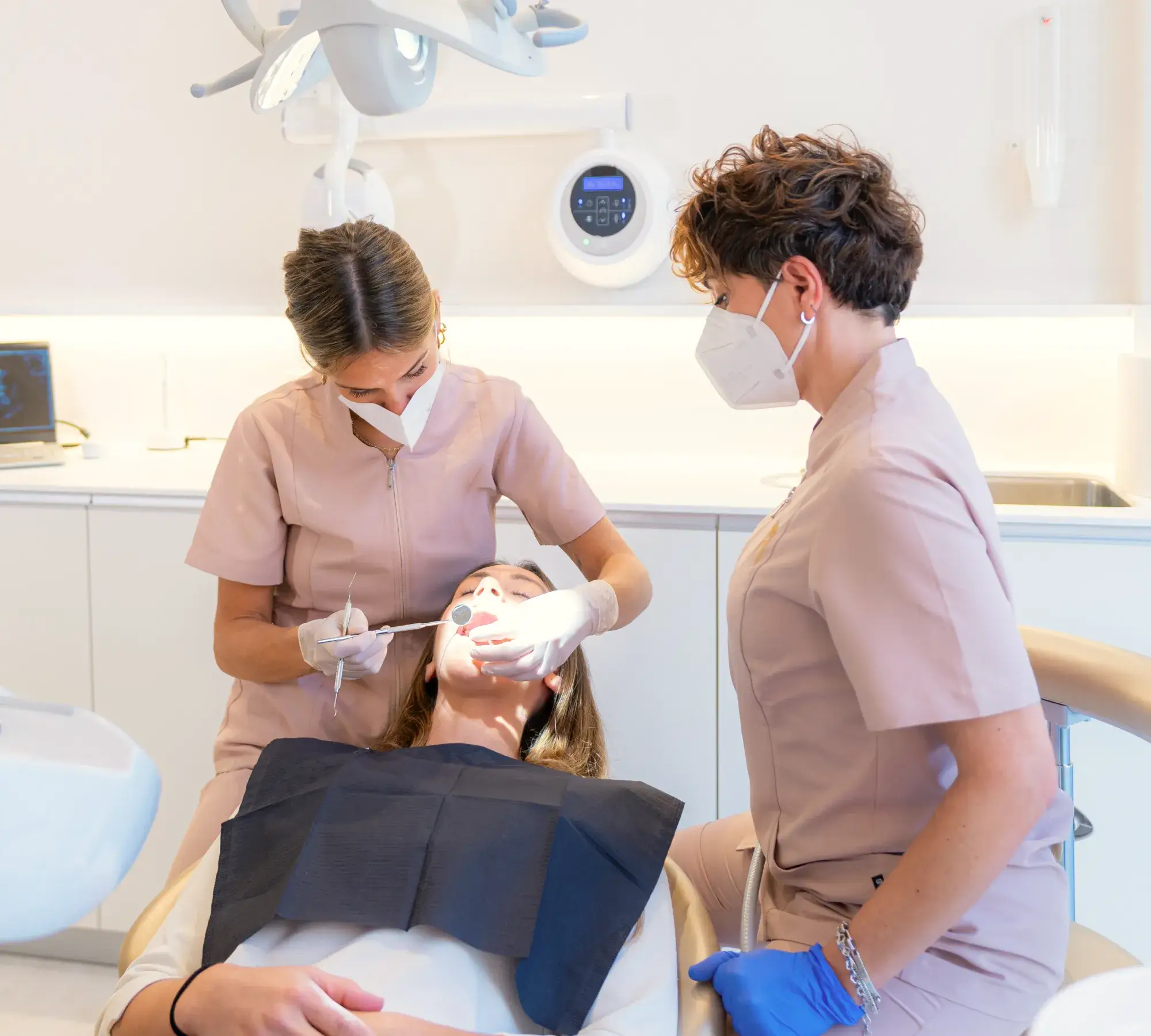Two dental professionals wearing masks and uniforms treating a patient reclining in a dental chair.