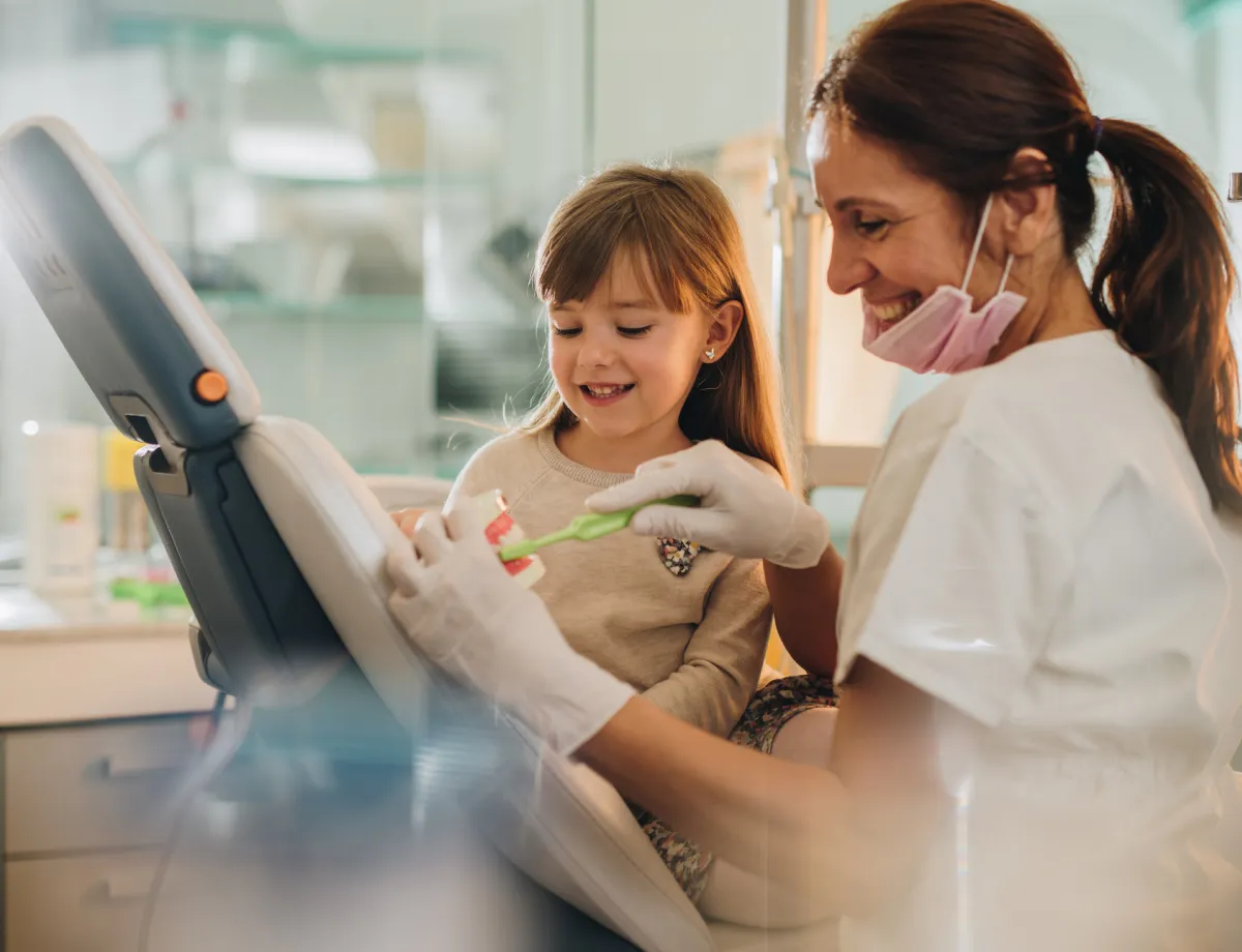 Female dentist demonstrating toothbrushing on a dental model to a smiling young girl in a dental office.