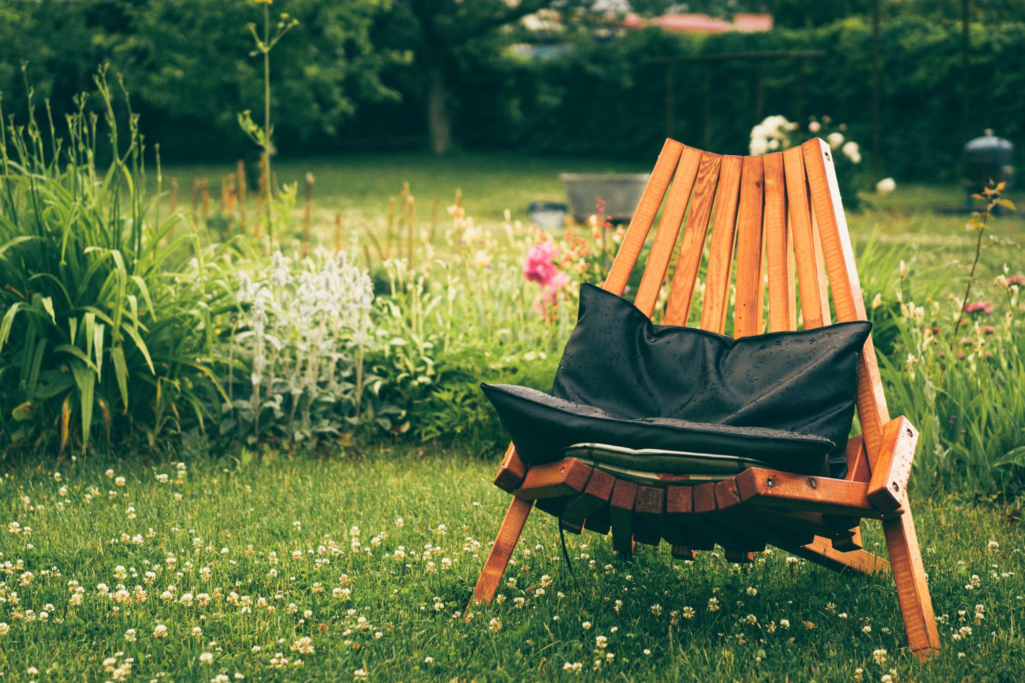 Wooden outdoor chair with black cushions placed on grass near a garden with various plants and flowers.