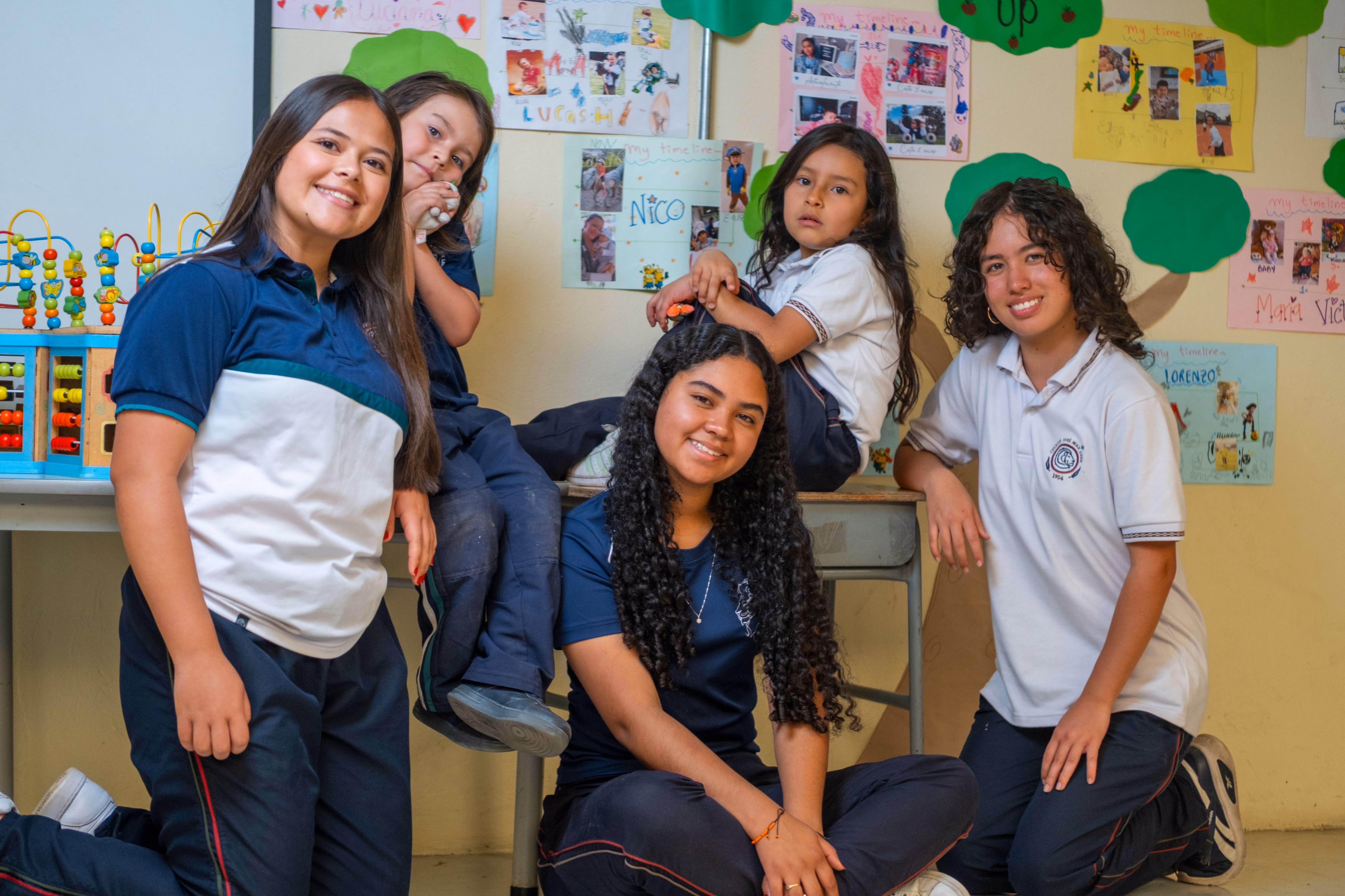 Cinco niñas en uniforme escolar posando sonrientes en un aula con dibujos en la pared y juguetes educativos.