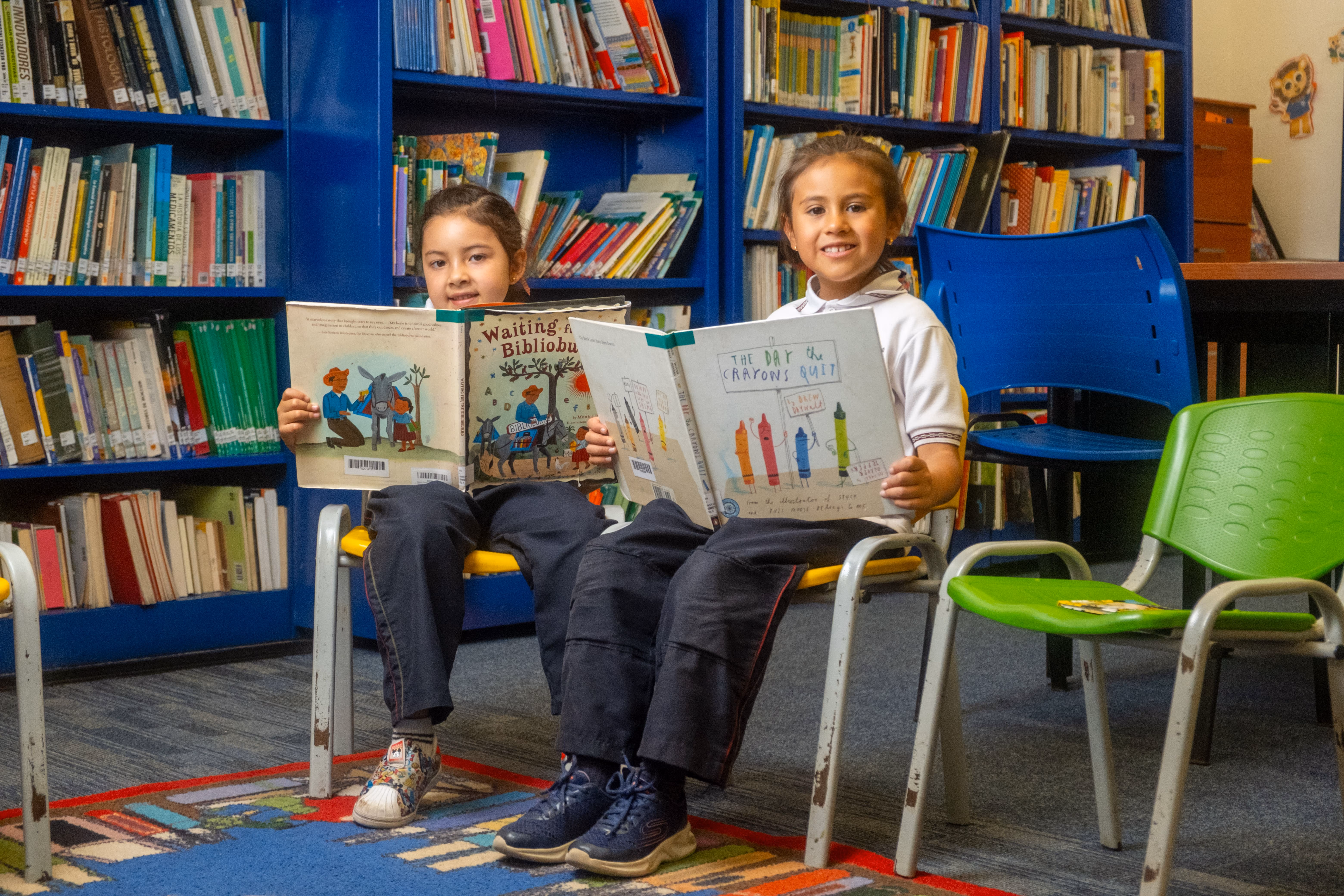 Dos niñas sonrientes sentadas en sillas en una biblioteca leyendo libros ilustrados.